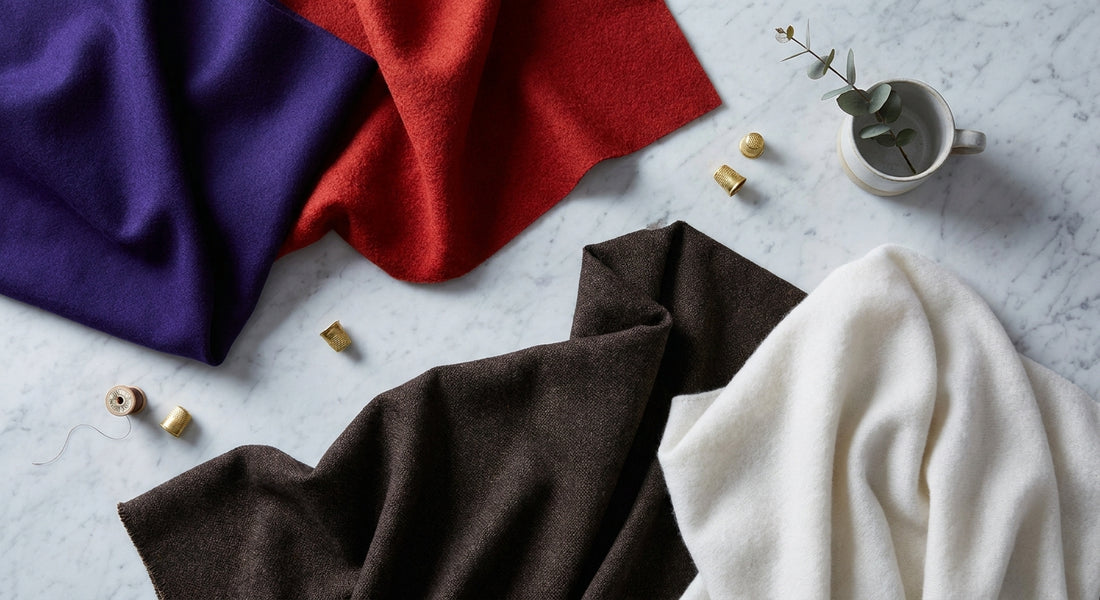 A flat-lay arrangement of premium outerwear fabric swatches in FW2026 trend colors including deep purple, chili red, espresso brown, and cloud white, draped across a cool marble surface under soft studio lighting.