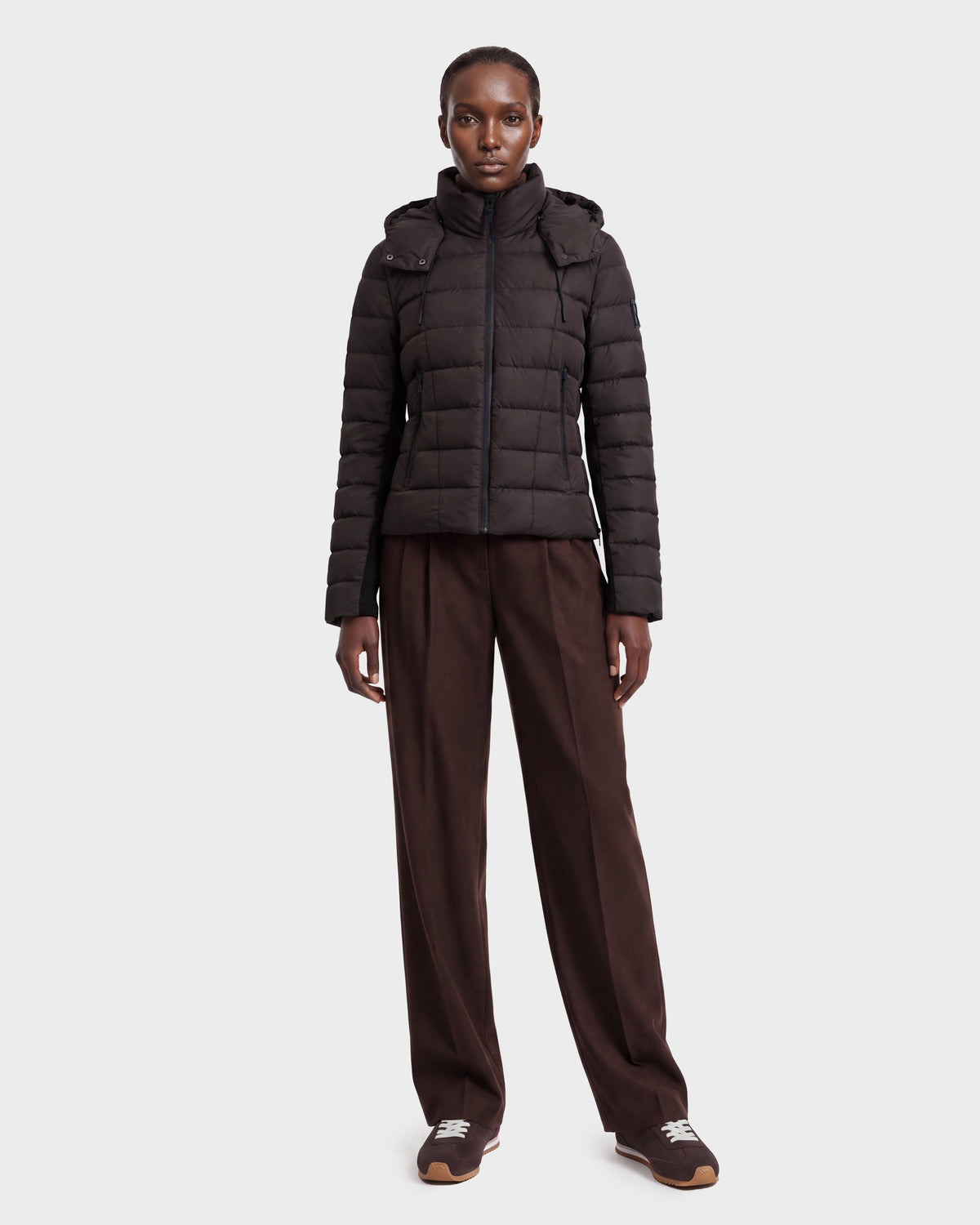 A person stands facing forward against a plain light gray background, wearing the Rudsak MELISSA - 8124824 Espresso/Black women’s lightweight down puffer. She pairs it with brown wide-leg pants and brown sneakers.