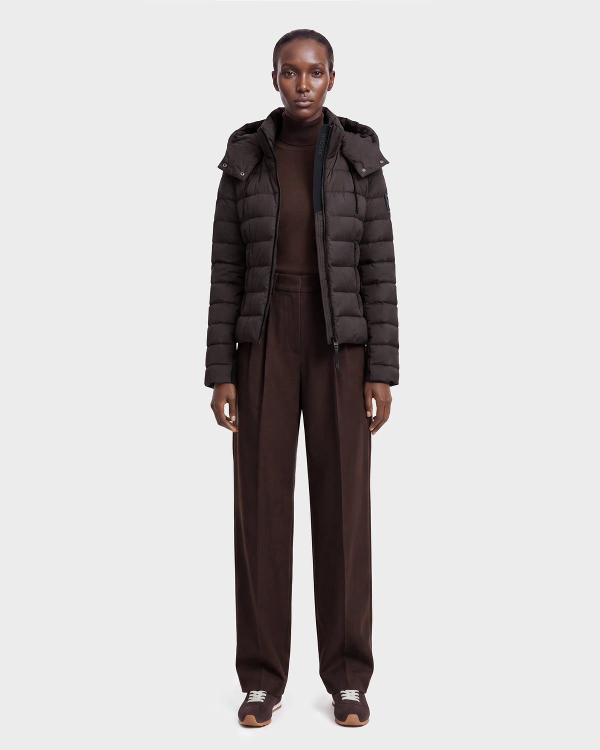 A person stands facing forward against a plain light gray background, wearing the Rudsak MELISSA - 8124824 Espresso/Black weatherproof down jacket, a dark brown turtleneck sweater, wide-leg brown pants, and sneakers.