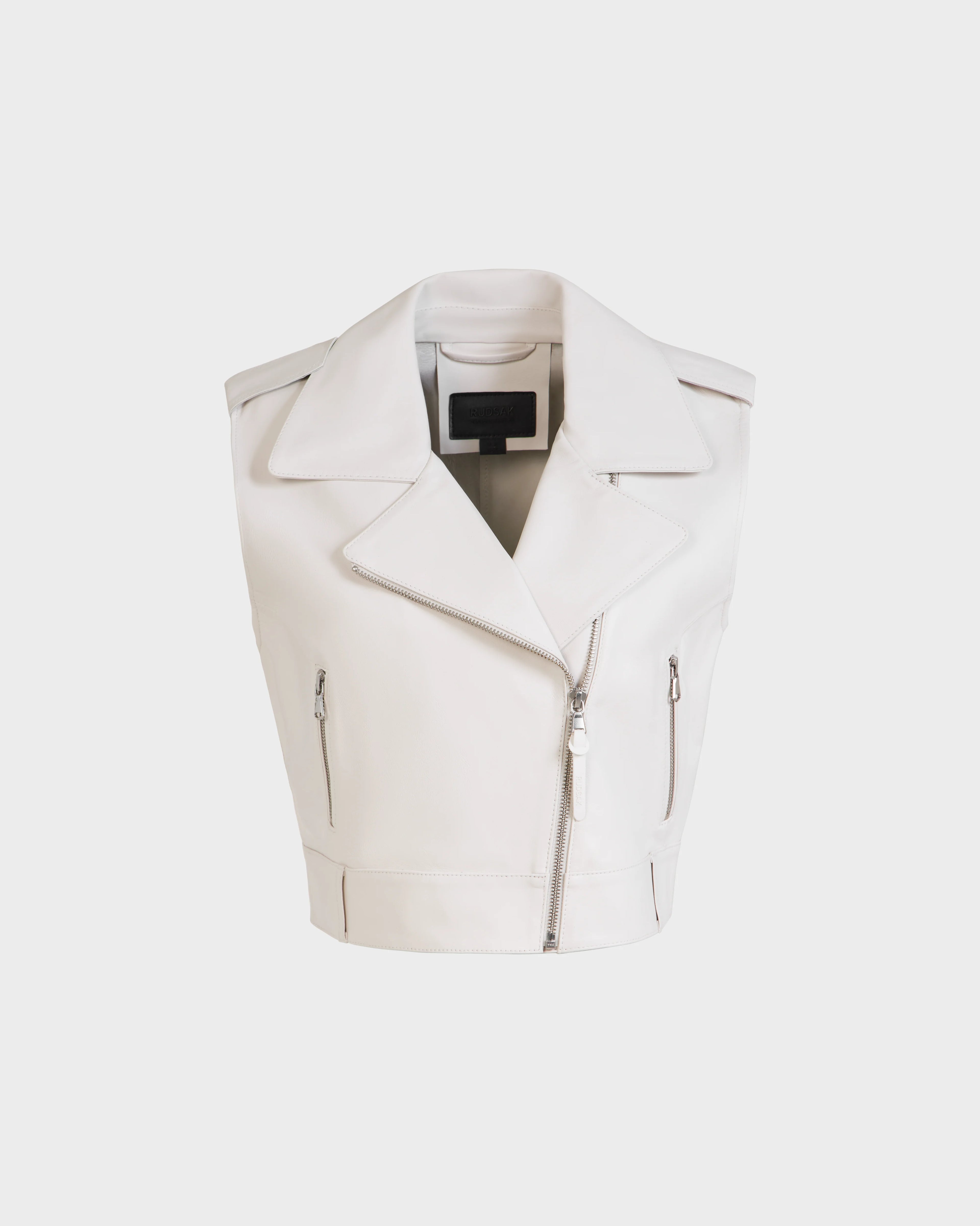 The Rudsak BETTY - 8125000 Optic White is a sleeveless cropped leather vest featuring wide lapels, an asymmetrical front zipper, and two zippered side pockets, shown on a plain white background.