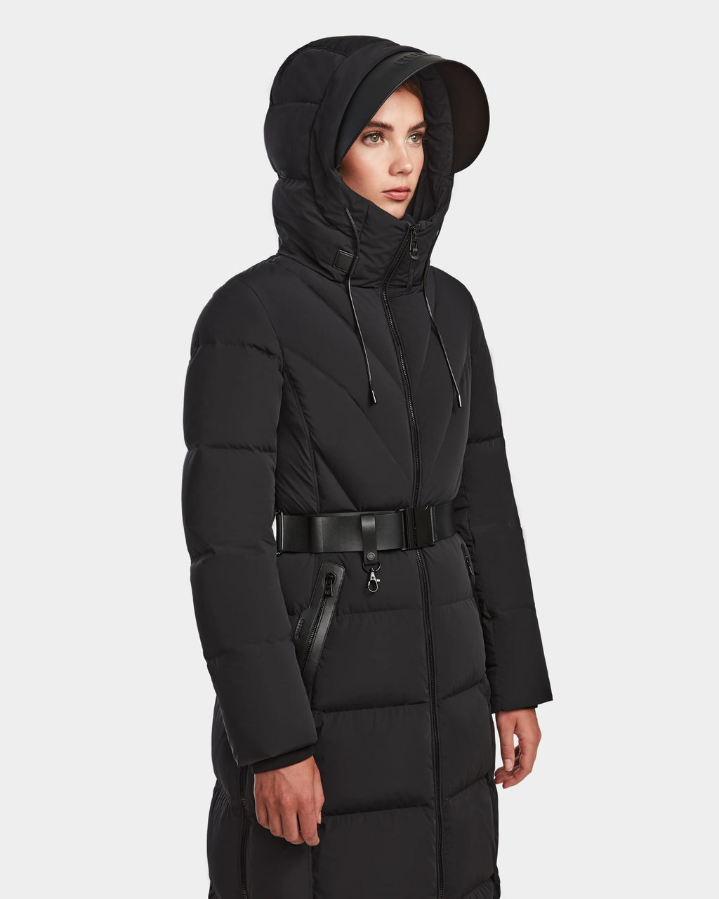 A woman models the Rudsak DALSA - 8124691 Black womens long-length down puffer coat with a hood, horizontal quilting, and zippered pockets against a light background, highlighting its stylish warmth and comfort.