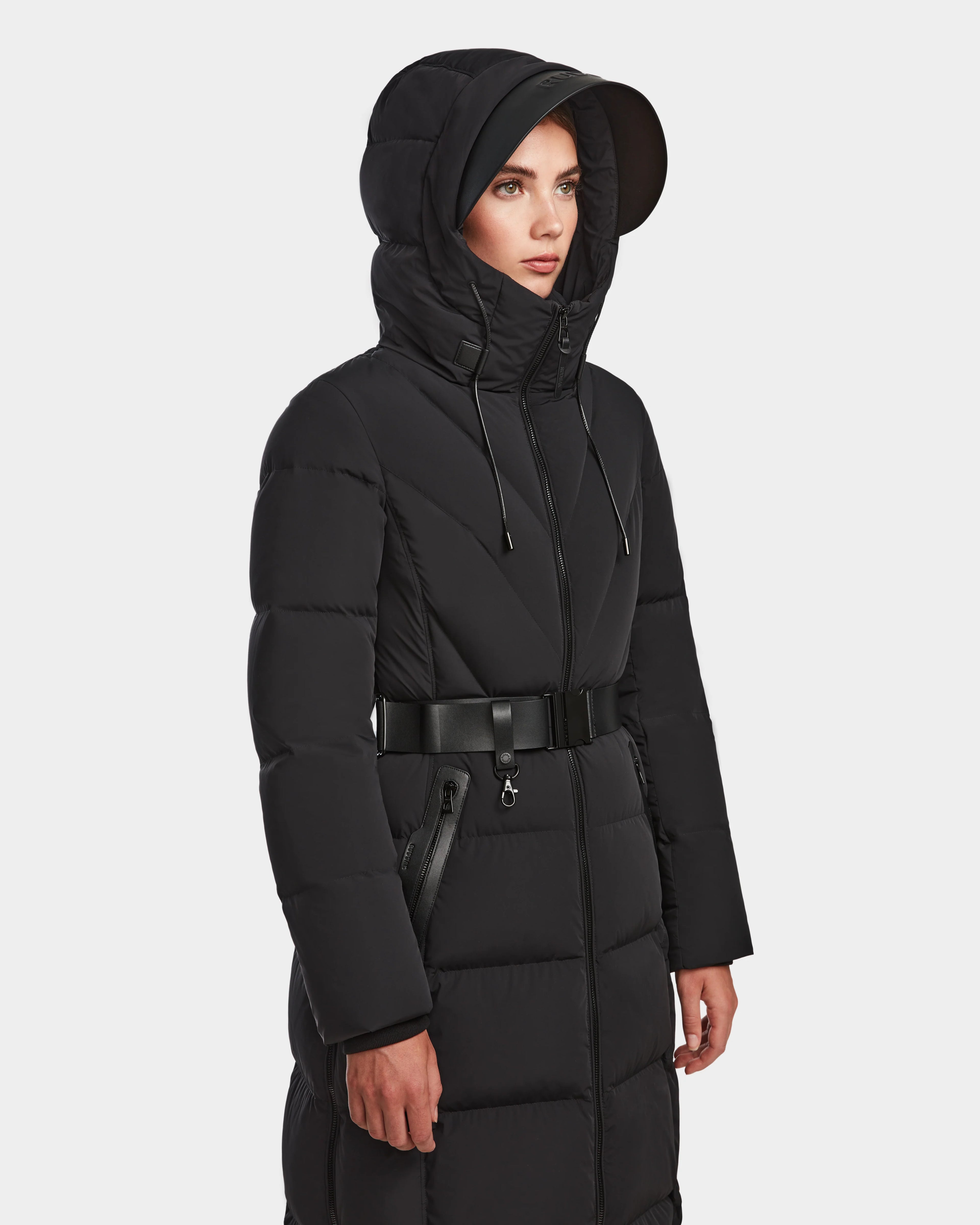 A woman models the Rudsak DALSA - 8124691 Black womens long-length down puffer coat with a hood, horizontal quilting, and zippered pockets against a light background, highlighting its stylish warmth and comfort.