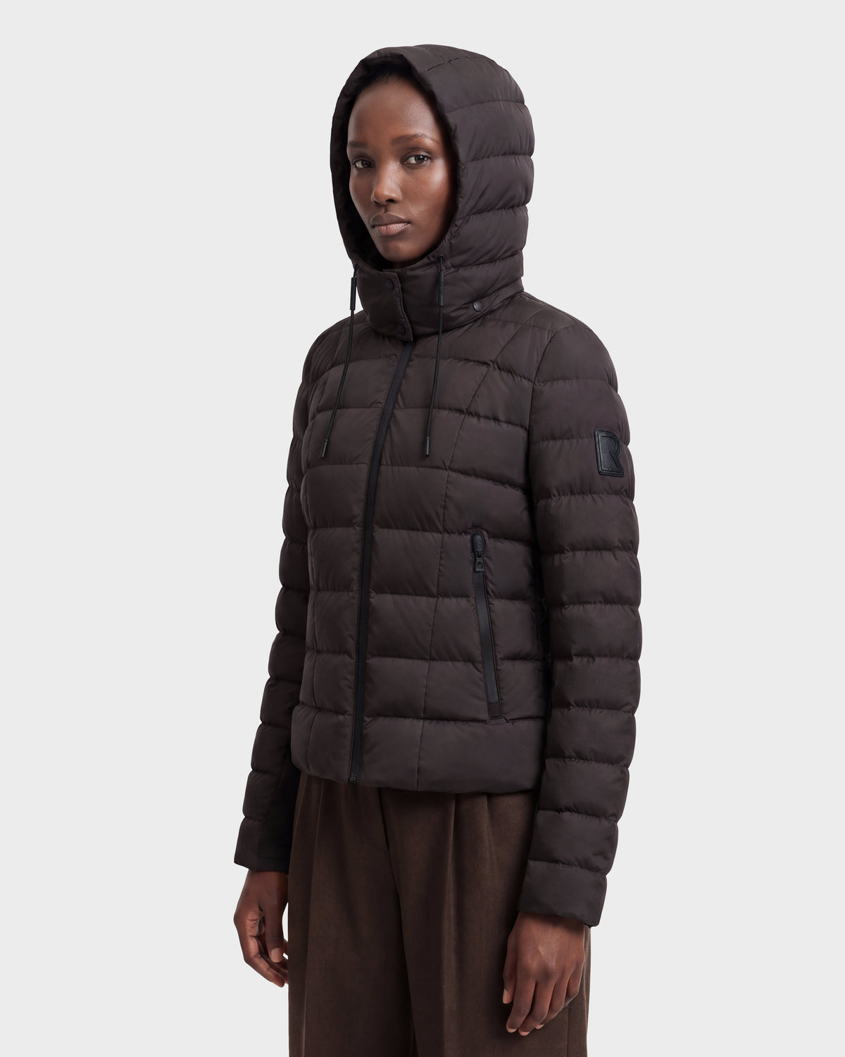 A person wearing the Rudsak MELISSA - 8124824 Espresso/Black women’s lightweight down puffer with a hood, made from RDS certified down, stands against a plain light background, gazing slightly to the side with a neutral expression.