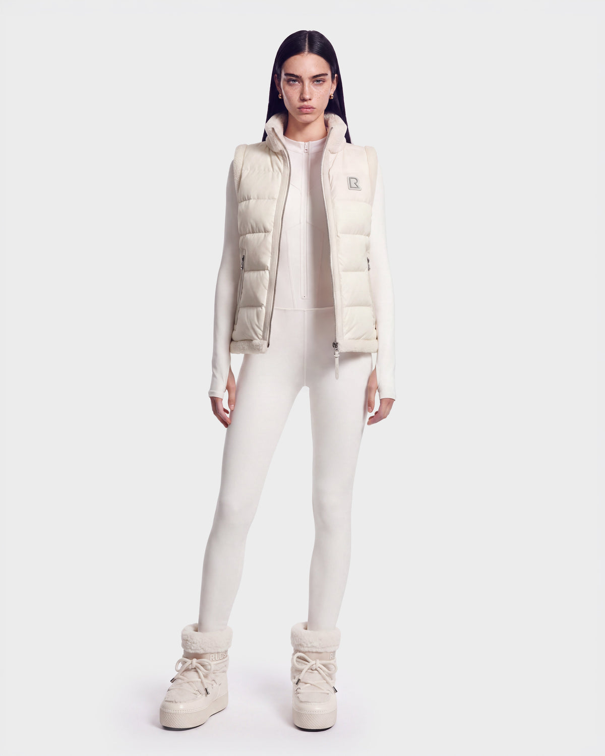 A woman stands facing forward, wearing a Rudsak HALI - 8126771 Vanilla puffer vest with down insulation over a fitted jumpsuit and high platform boots, set against a plain white background.