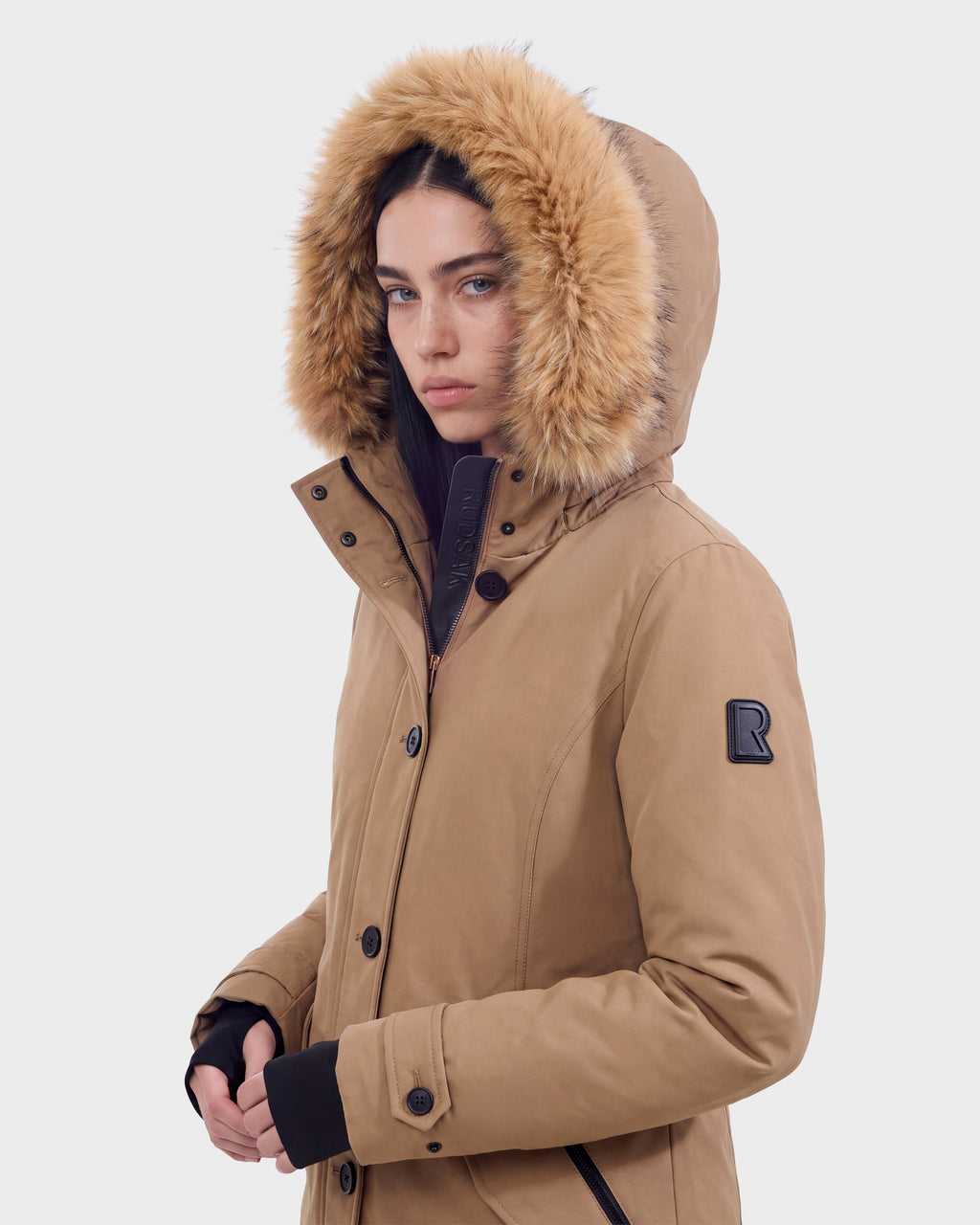 A person wearing the Rudsak KEYSHIA - 8126517 Camel down winter coat, featuring a fur-lined hood and RDS-certified filling, stands against a plain background, looking slightly to the side with a neutral expression.