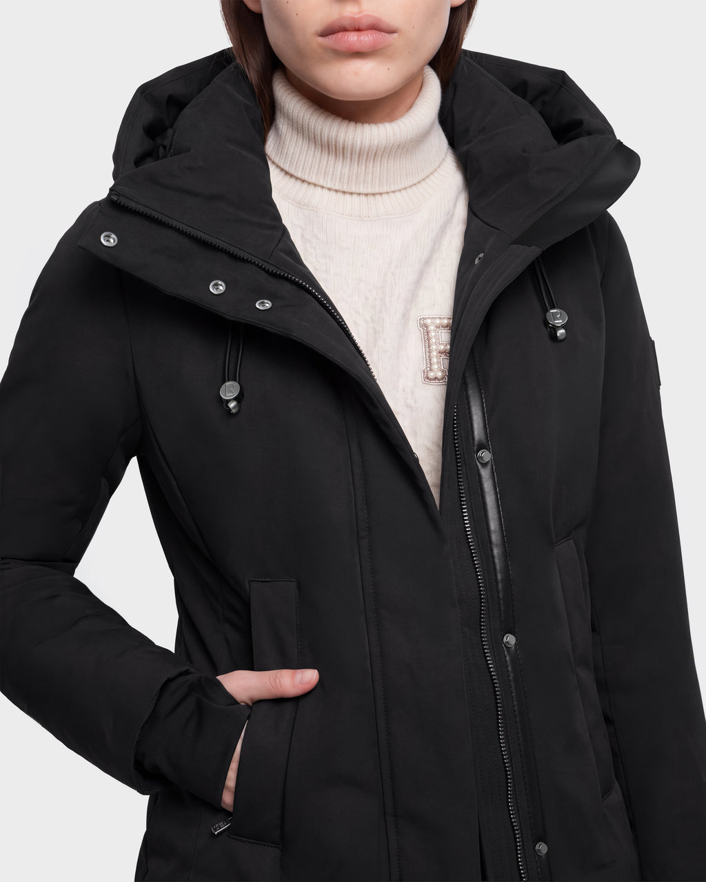 A person wearing the Rudsak OPAL - 8126618 Black hooded winter coat over a cream turtleneck sweater with an embroidered letter E, standing with one hand in the coat pocket.