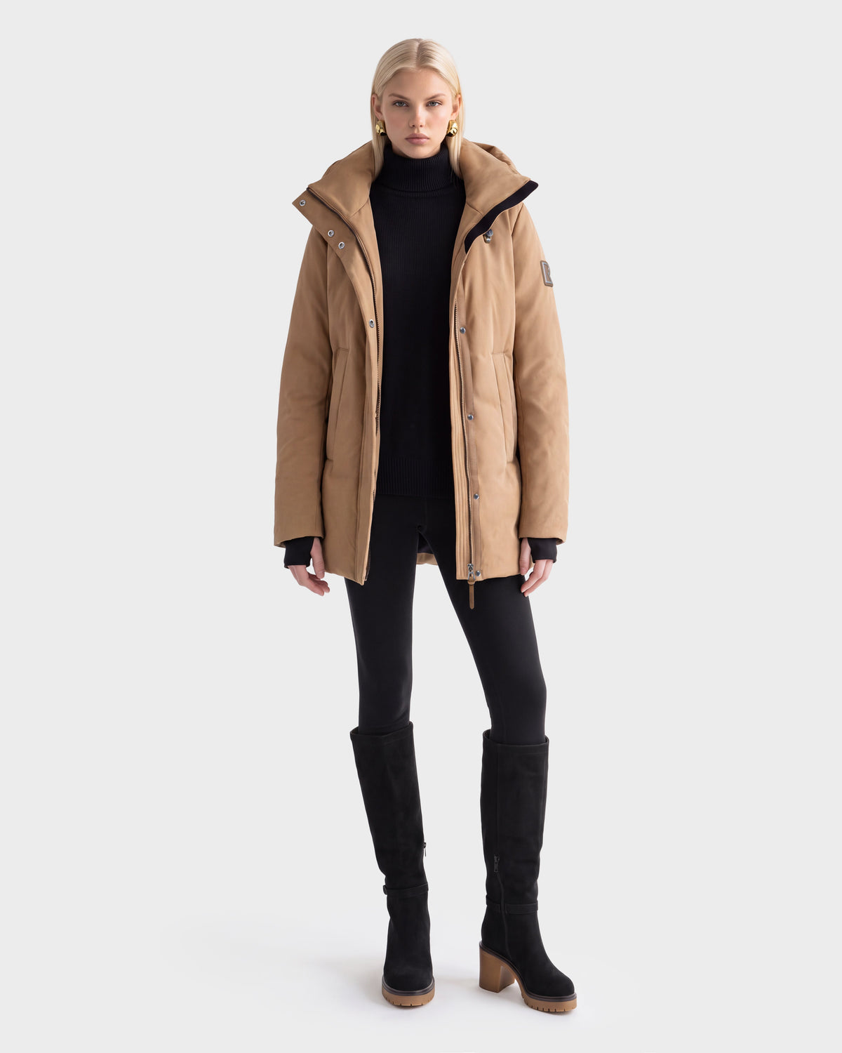 A person wears the Rudsak OPAL - 8126618 Camel winter coat, black turtleneck, black leggings, and knee-high boots while standing against a plain white background.