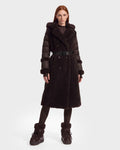 A woman with long red hair is seen wearing the NOELLE - 8126971 Espresso belted coat by Rudsak, styled as a puffer. She pairs it with black leggings and black fur-trimmed boots, standing against a plain white background.