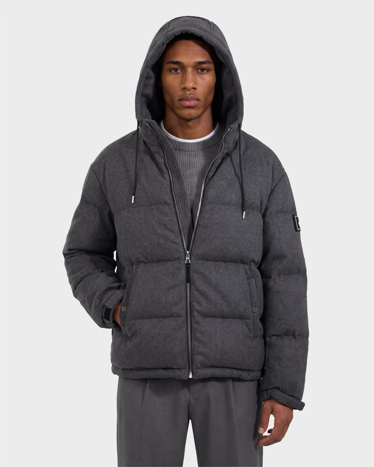 A person is seen wearing the Rudsak STERLING W - 6126760 Heather Charcoal puffer jacket with a hood, paired with gray pants and a ribbed gray sweater. The jacket is zipped up, and the individual has one hand tucked into a pocket, standing against a plain light background.