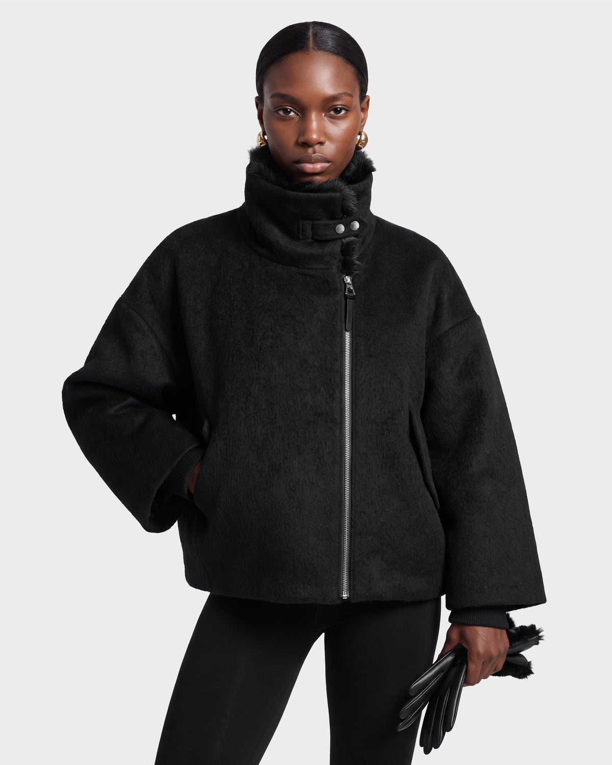 A woman models the Rudsak ZARIA - 8126999 Black high-collar jacket and matching leggings, holding black gloves. She wears gold earrings and maintains a serious expression while posing against a plain light gray background.
