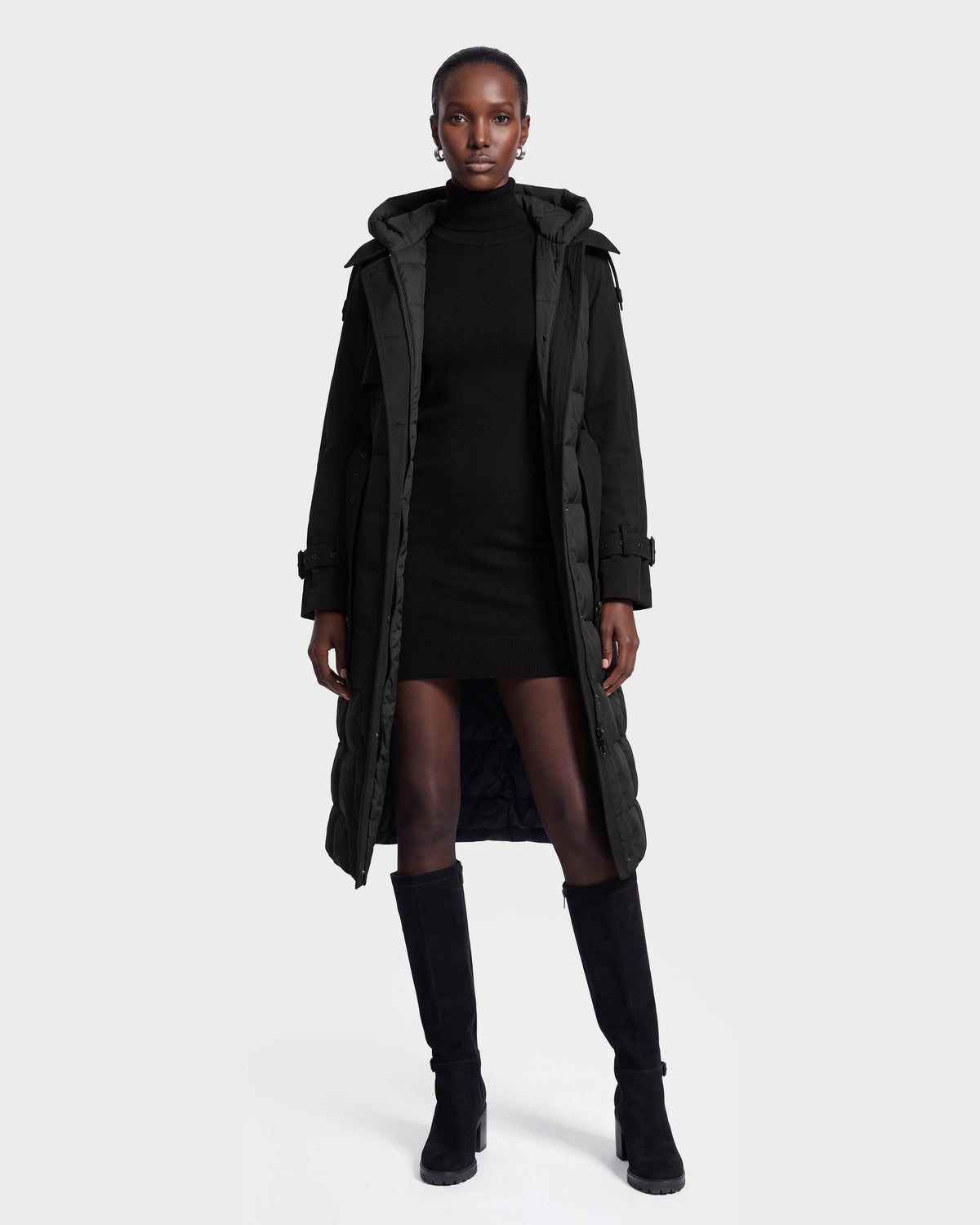 A woman stands facing forward, wearing the Rudsak ELVIE - 8126512 Black long puffer coat over a black turtleneck dress and knee-high black boots, against a plain white background.