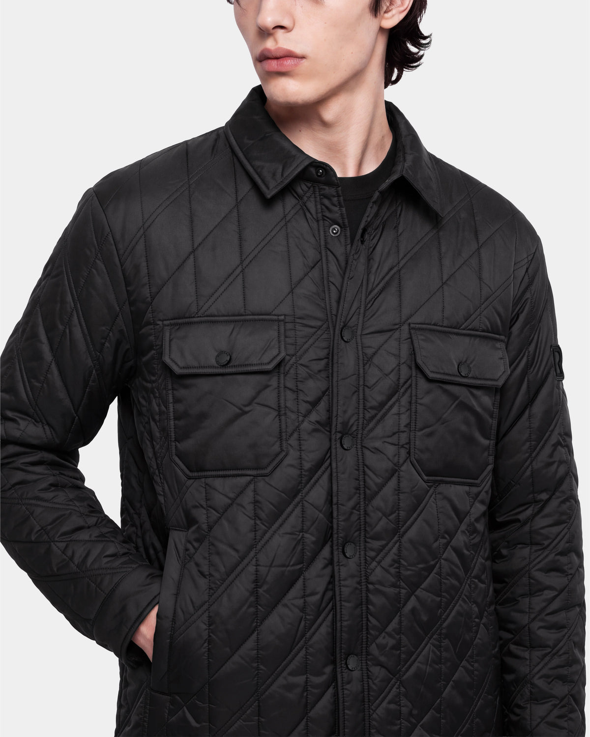 A person wears the Rudsak GRIFFIN - 6126758 Black jacket, a quilted style featuring snap buttons and two chest pockets, standing with one hand in a side pocket. Their face is partially visible as they look to the side.