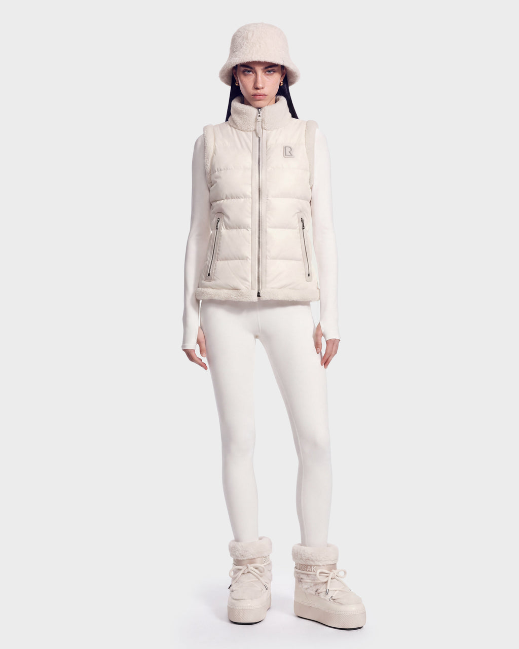 A person stands against a plain light background wearing an all-white ensemble, highlighted by the Rudsak HALI - 8126771 Vanilla puffer vest with down insulation, paired with a bucket hat, long-sleeve top, leggings, and winter boots.