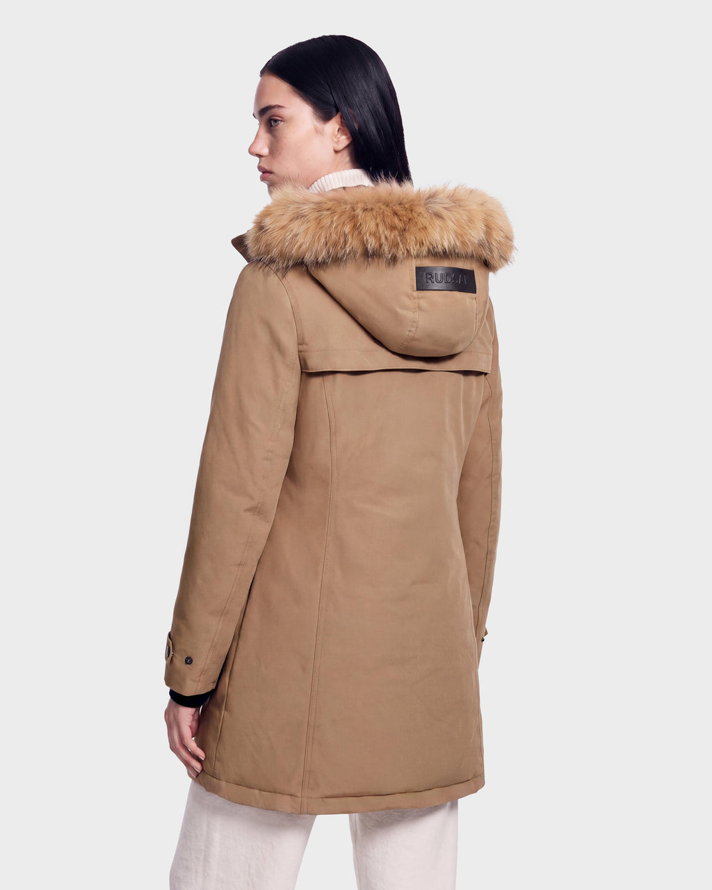A person with dark straight hair is seen from the back wearing the Rudsak KEYSHIA - 8126517 Camel women’s down parka, which features a fur-lined hood, an RDS-certified down fill, and a black patch on the upper back. The winter coat is paired with cream-colored pants.