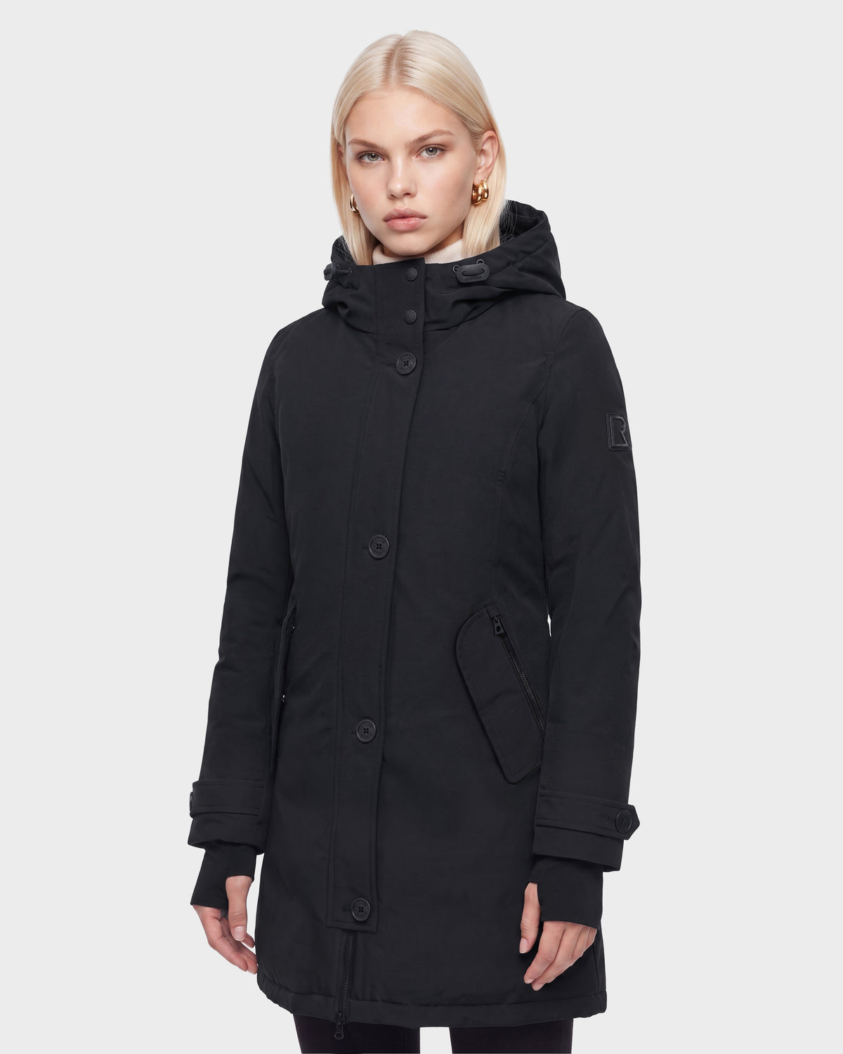 A woman with blonde hair wears the Rudsak KEYSHIA - 8126517 Black long down winter coat, featuring large buttons, side pockets, and a high collar, against a plain white background.