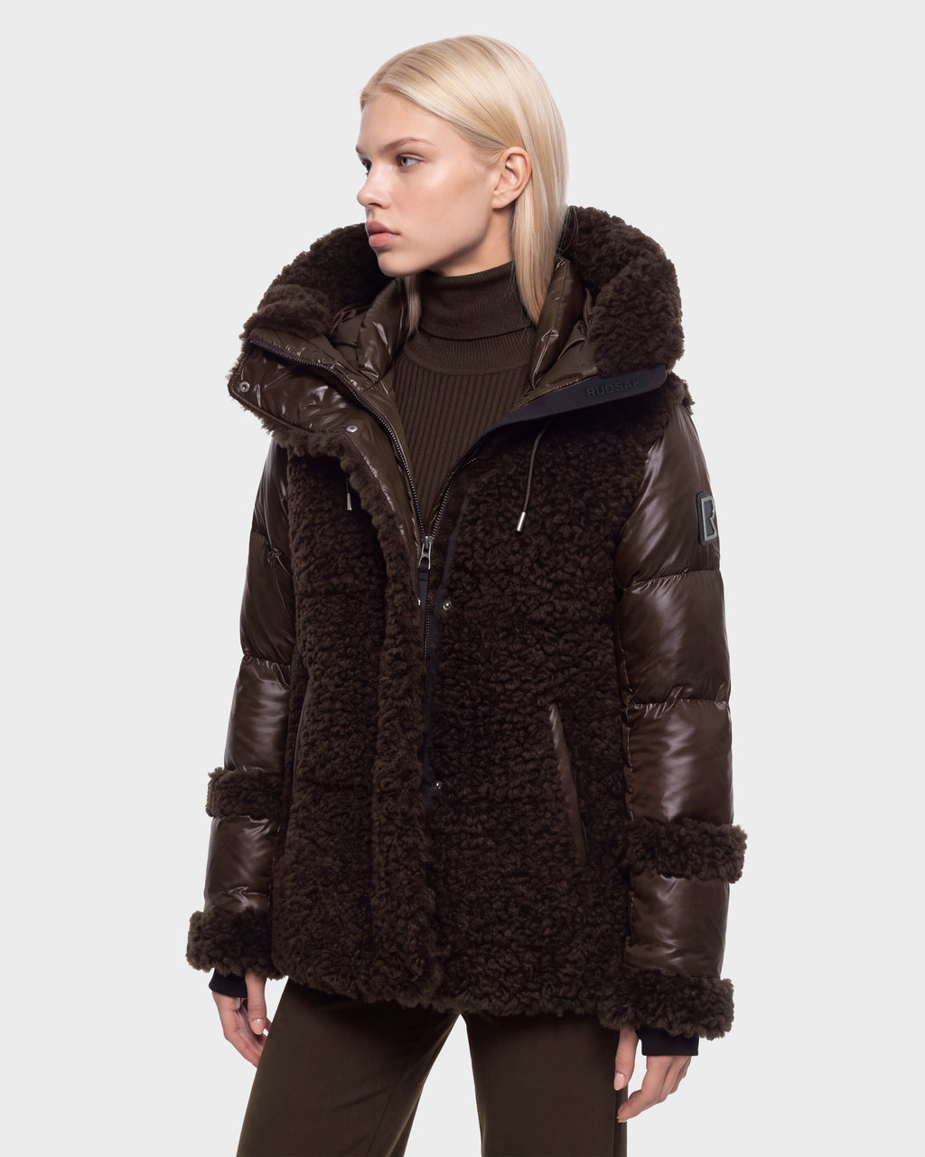 A person with straight blonde hair wears the Rudsak MARISSA - 8126728 Espresso dark brown plush mixed-texture winter jacket, reminiscent of a cozy sherpa style, layered over a turtleneck. They are standing against a plain light background, looking to their left.