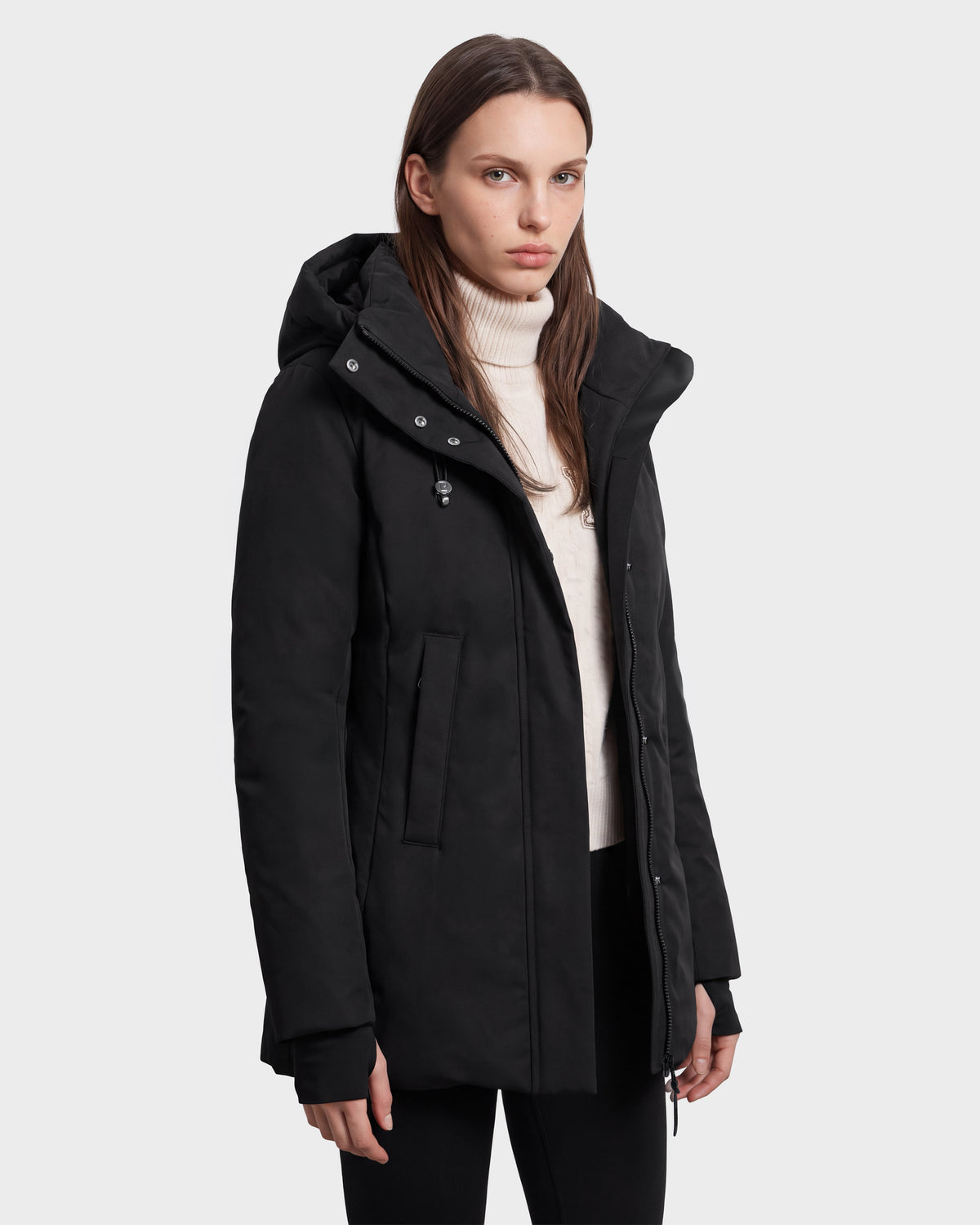 A woman wearing the Rudsak OPAL - 8126618 Black hooded winter coat over a white turtleneck sweater stands against a plain light background, looking at the camera with a neutral expression.