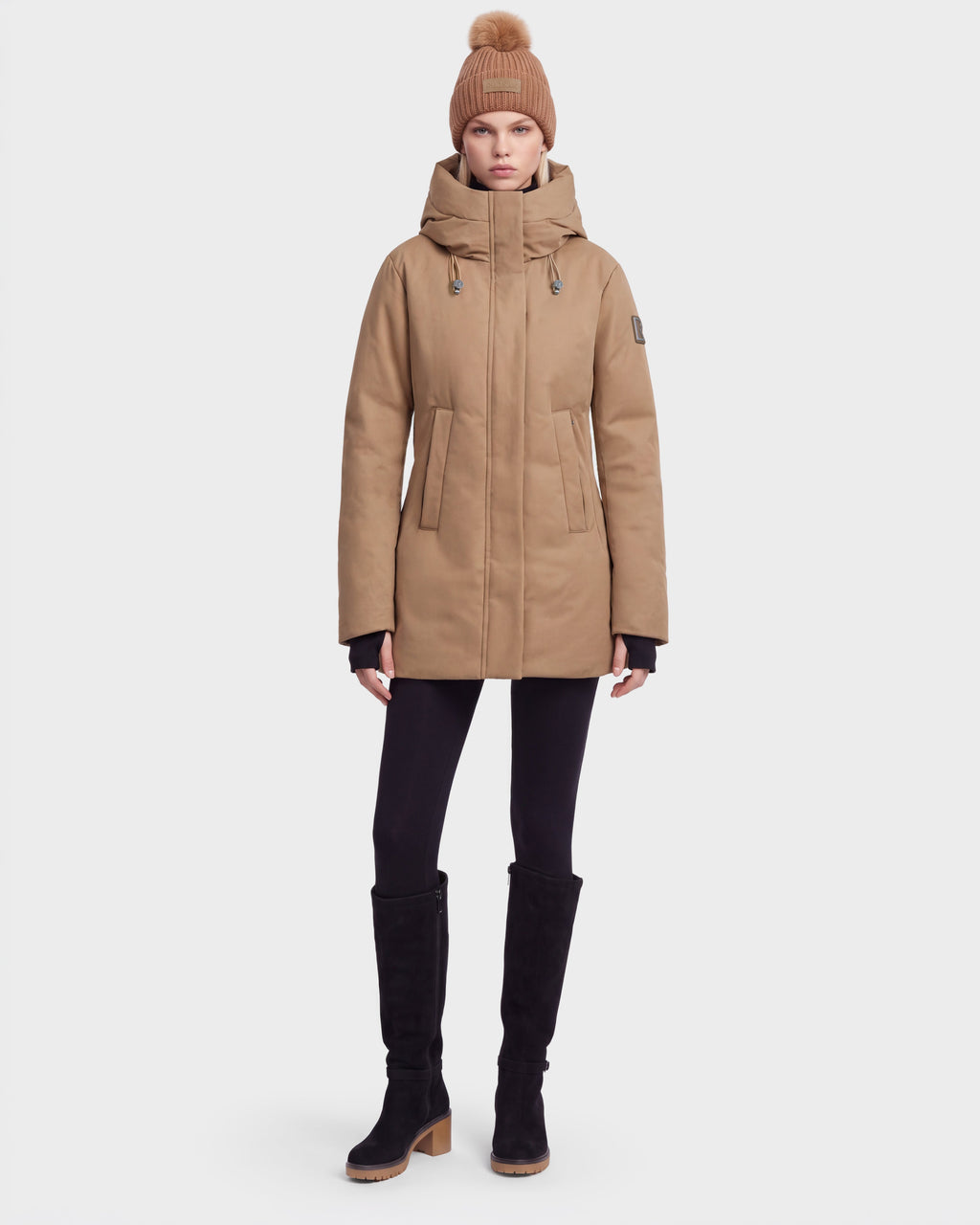 A woman in the Rudsak OPAL - 8126618 Camel winter coat, a matching pom-pom knit hat, black leggings, and knee-high black boots stands facing forward against a plain light background.