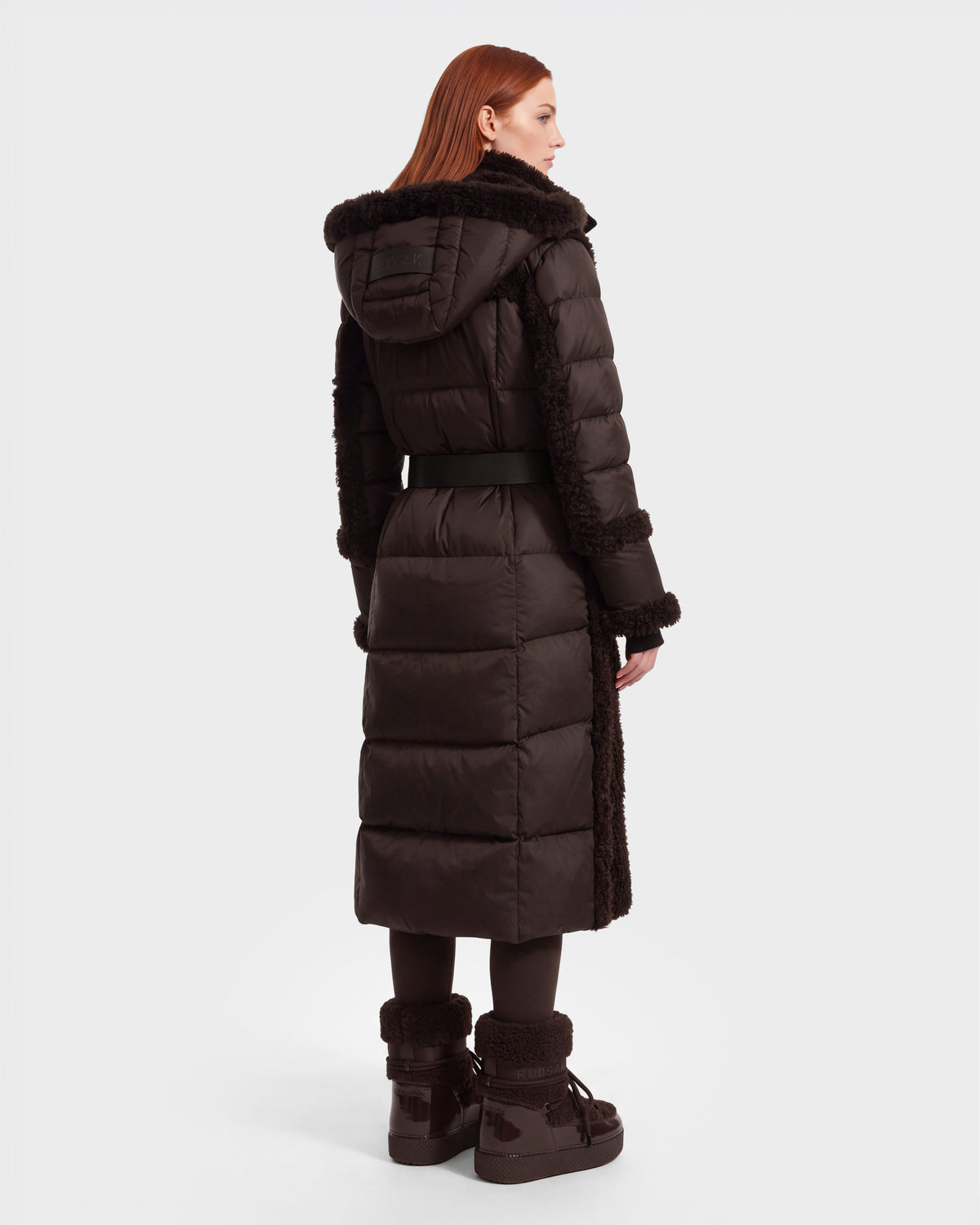 A woman with red hair is seen from the back against a plain white background, wearing the Rudsak NOELLE - 8126971 Espresso long, dark brown hooded winter coat with a fur-trimmed hood and matching boots.