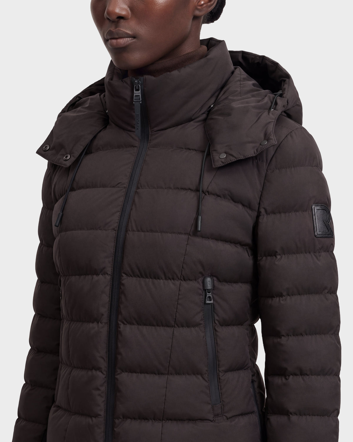 A person wearing the Rudsak MELISSA - 8124824 Espresso/Black women’s lightweight down puffer with a hood and zippered pockets, shown from the shoulders up against a light background.