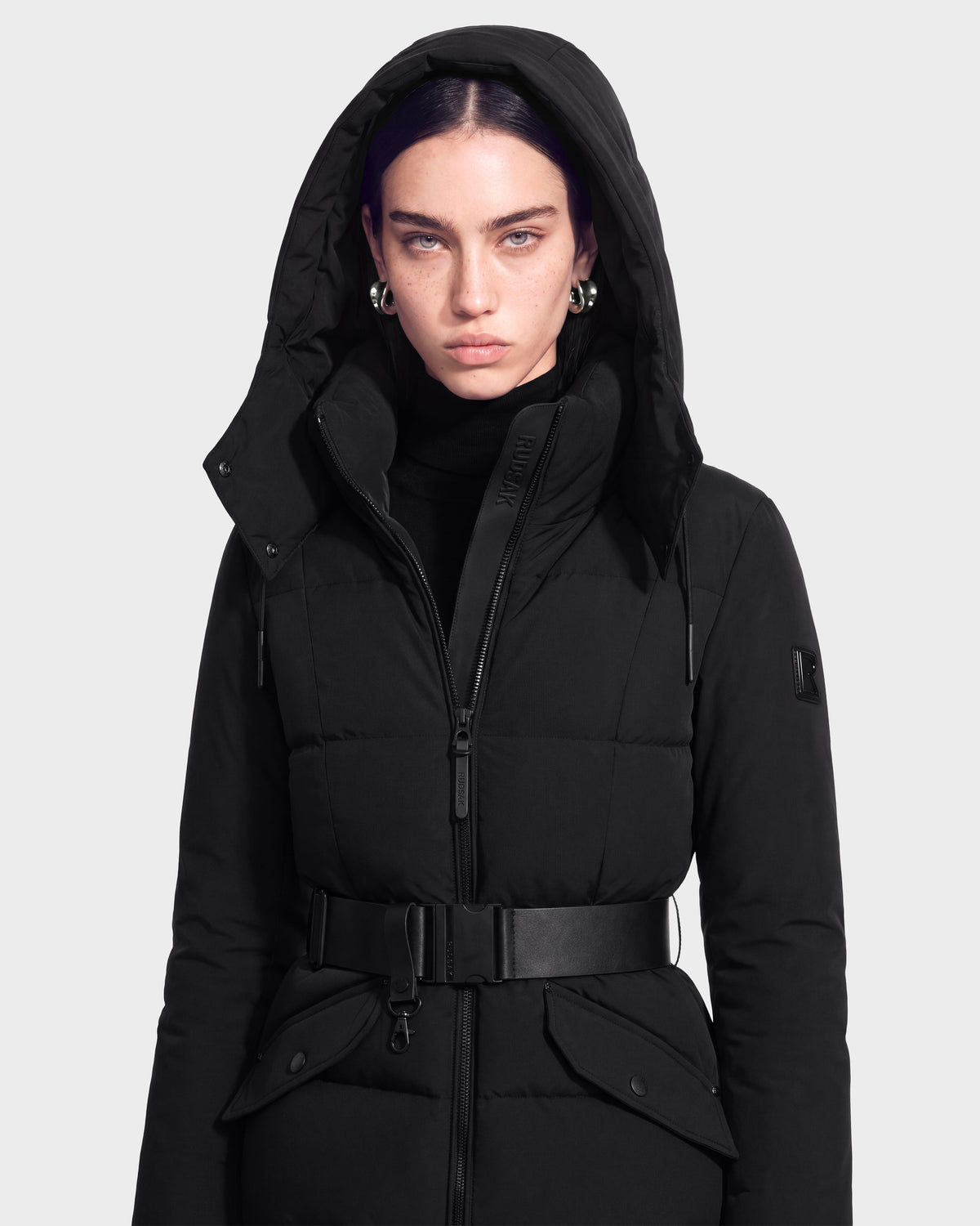 A person with straight dark hair, wearing the Rudsak ADELINE - 8124544 Black quilted winter jacket with a belt and silver hoop earrings, stands facing forward against a plain light background.