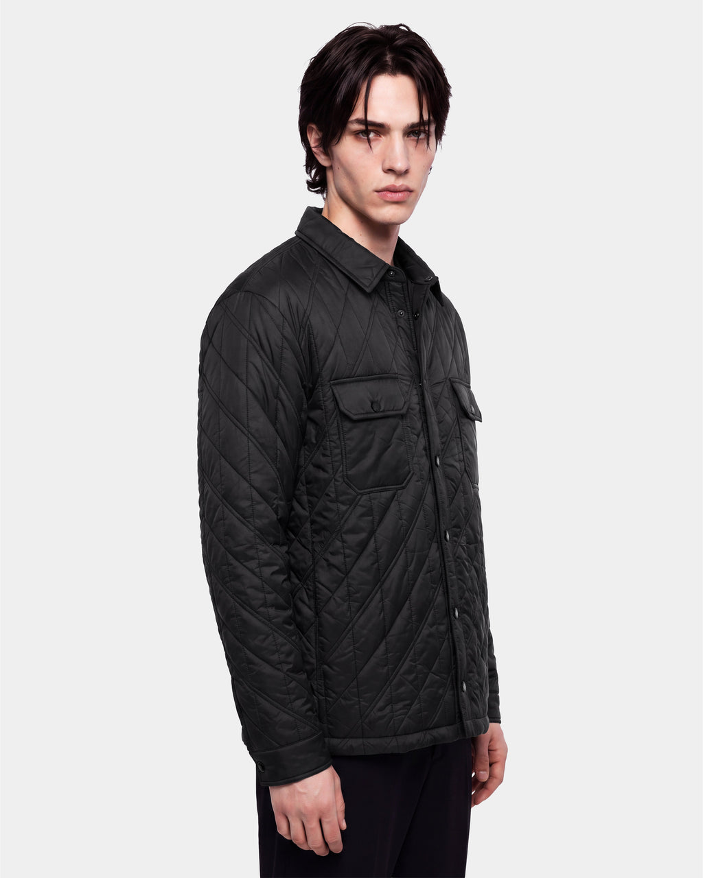 A man with dark hair wears the Rudsak GRIFFIN - 6126758 Black quilted jacket, featuring snap buttons and front chest pockets, standing against a plain white background.