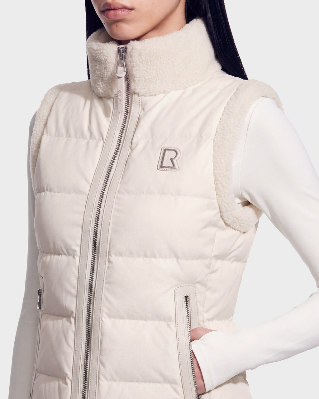 A woman wears the Rudsak HALI - 8126771 Vanilla sleeveless puffer vest with down insulation and a fuzzy collar over a white long-sleeve top, hand in pocket, featuring the stitched Rudsak logo on the chest.