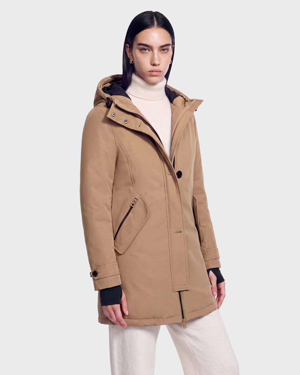 A person wearing the Rudsak KEYSHIA - 8126517 Camel women’s down parka over a white turtleneck sweater and light pants stands against a plain light gray background, looking directly at the camera. The winter coat features RDS-certified down for warmth and comfort.