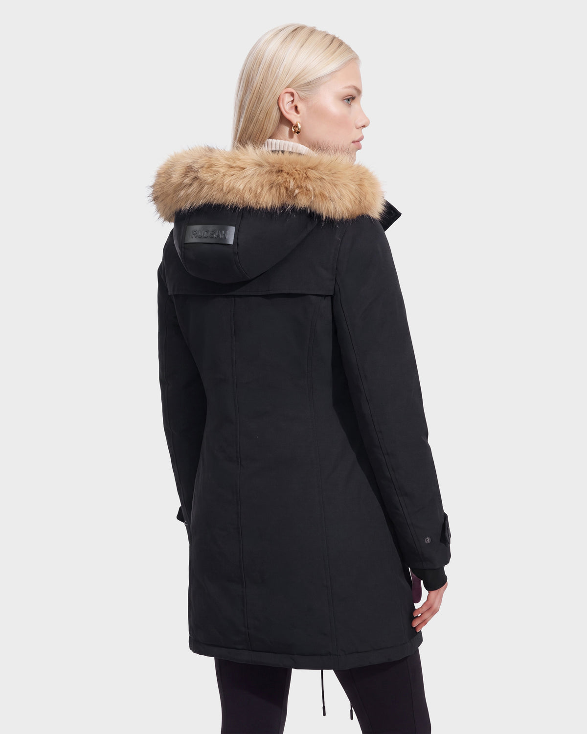 A woman with straight blonde hair is seen from behind wearing the Rudsak KEYSHIA - 8126517 Black women’s down parka with a tan fur-trimmed hood against a plain white background.