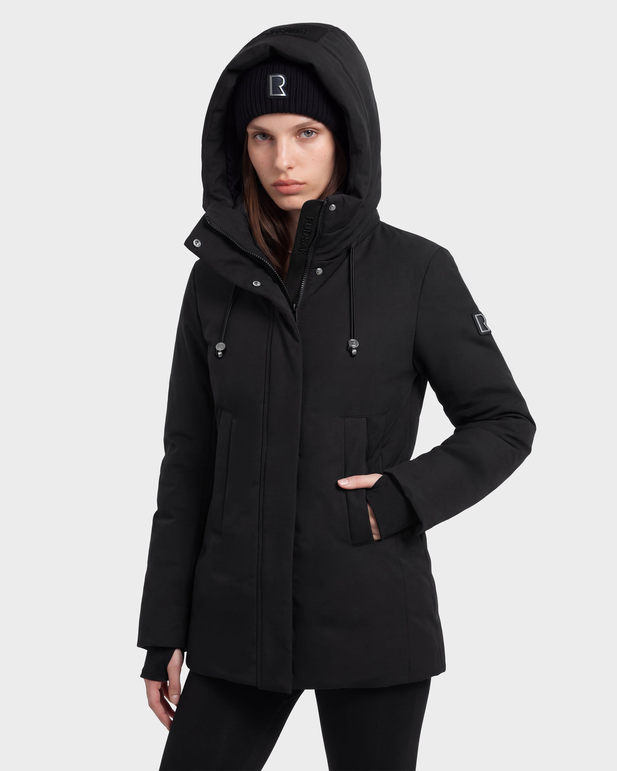 A person stands facing forward with hands in the pockets of a Rudsak OPAL - 8126618 Black hooded winter jacket and a matching black beanie, both featuring a small white logo, against a plain light background.