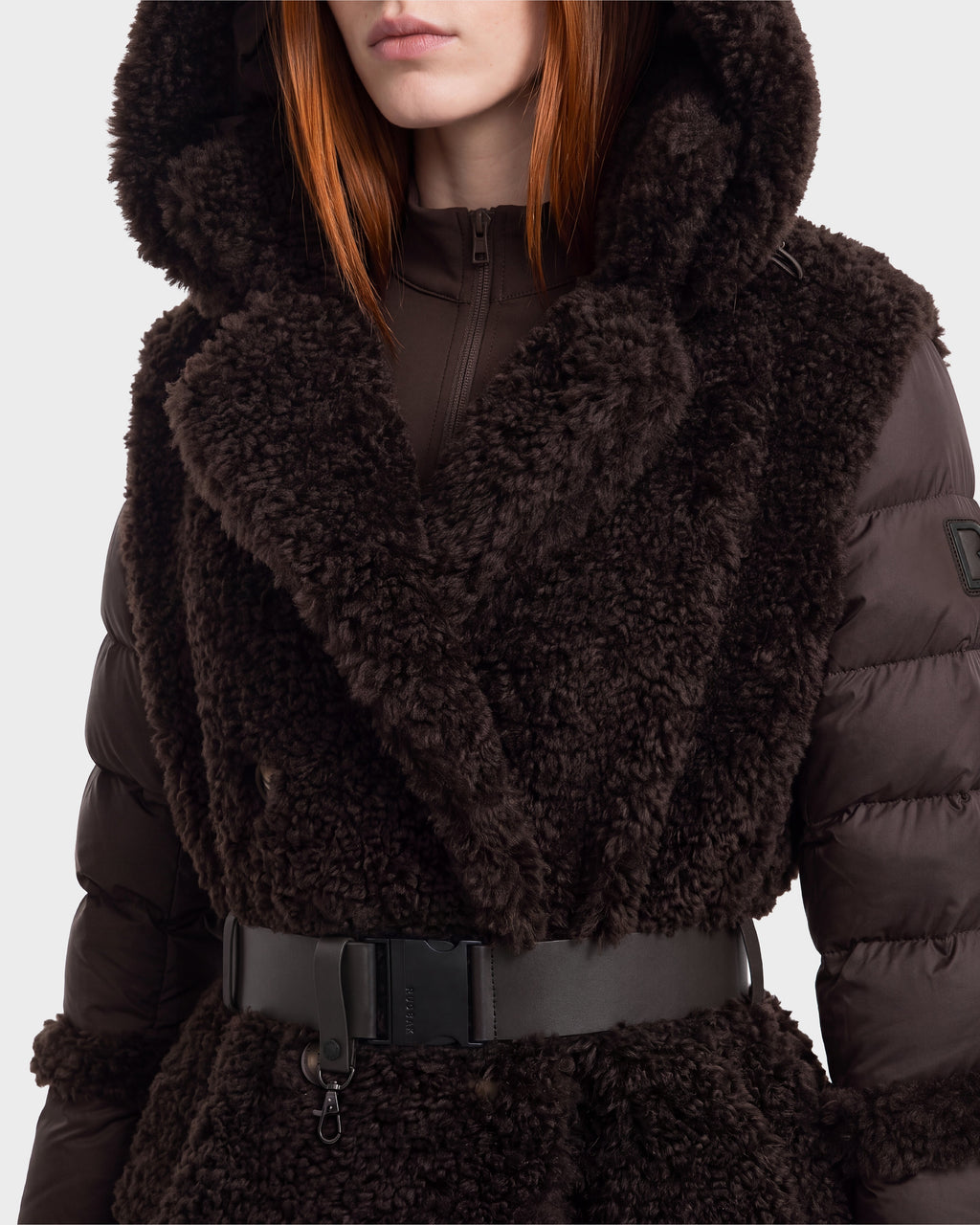 A person is seen wearing the NOELLE - 8126971 Espresso winter coat by Rudsak, featuring a dark brown, textured finish with a plush faux fur style. The coat includes a wide collar, puffy sleeves, hood, and is cinched at the waist with a black belt. Only the lower part of their face is visible, highlighting the luxurious texture and cozy design of this statement piece.