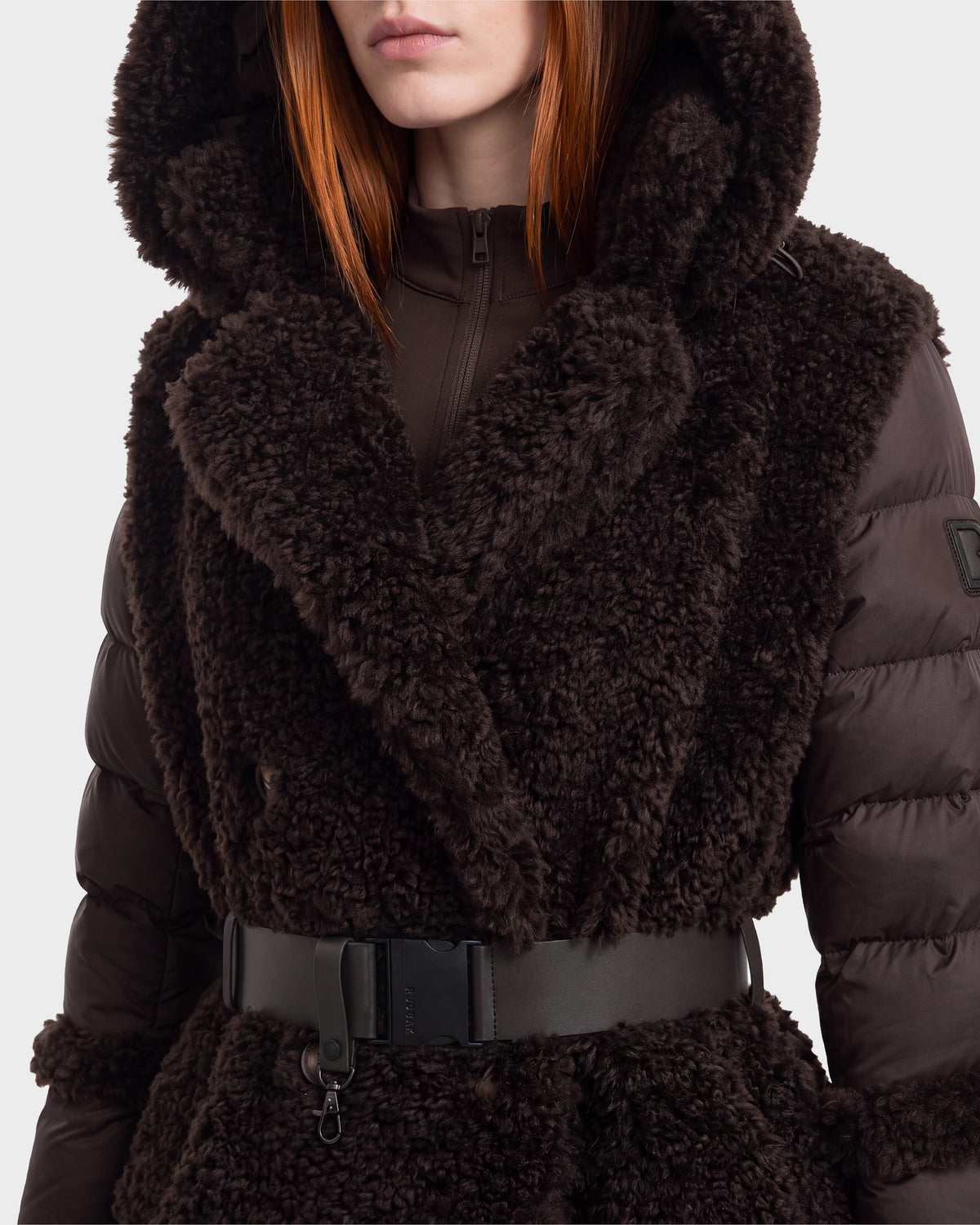 A person is seen wearing the NOELLE - 8126971 Espresso winter coat by Rudsak, featuring a dark brown, textured finish with a plush faux fur style. The coat includes a wide collar, puffy sleeves, hood, and is cinched at the waist with a black belt. Only the lower part of their face is visible, highlighting the luxurious texture and cozy design of this statement piece.