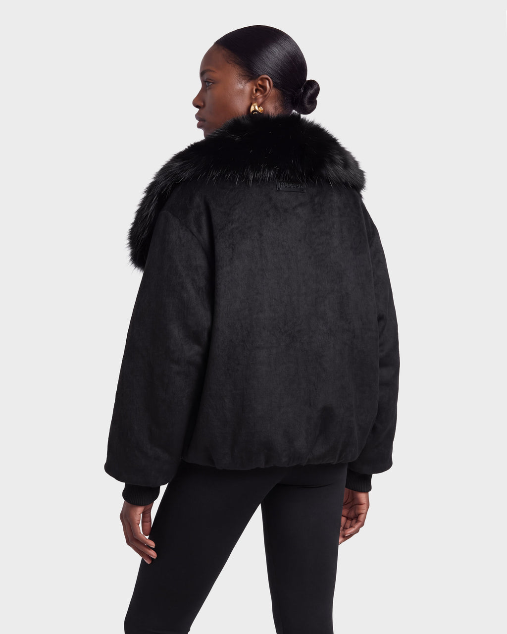 A woman stands facing away, wearing the Rudsak ZARIA - 8126999 Black jacket with a faux fur collar and black pants against a plain white background.