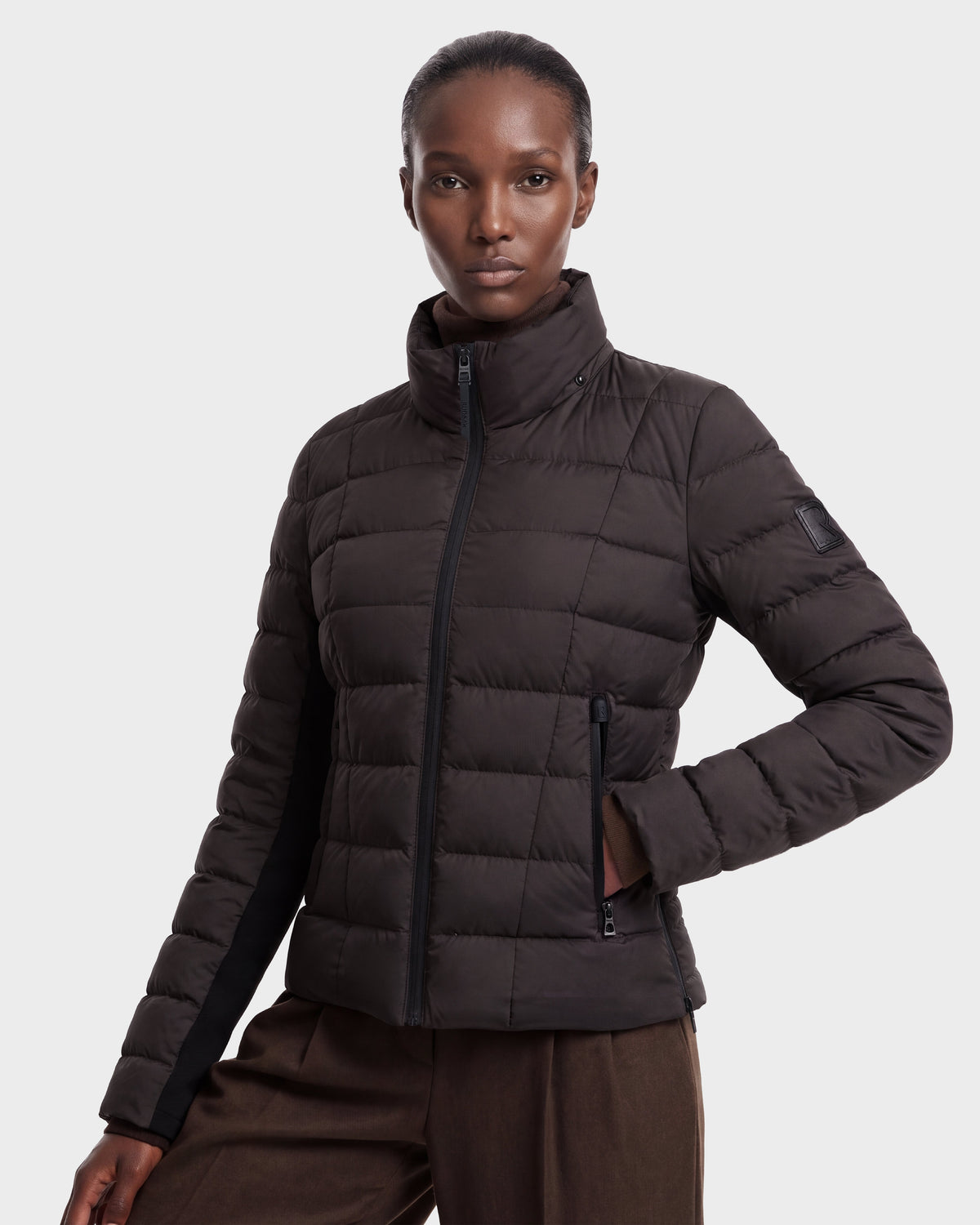 A woman wearing the Rudsak MELISSA - 8124824 lightweight down puffer in Espresso/Black poses with her left hand in her pocket against a plain light background.