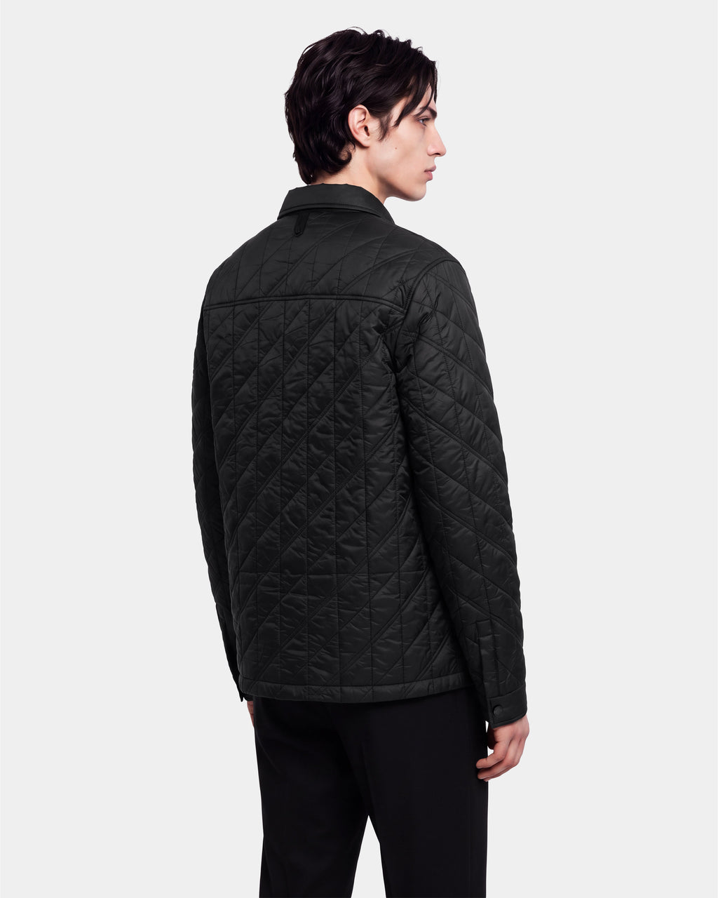 A person with dark hair is standing and facing away, looking to the side against a plain light background, wearing the Rudsak GRIFFIN - 6126758 Black quilted jacket and black pants.