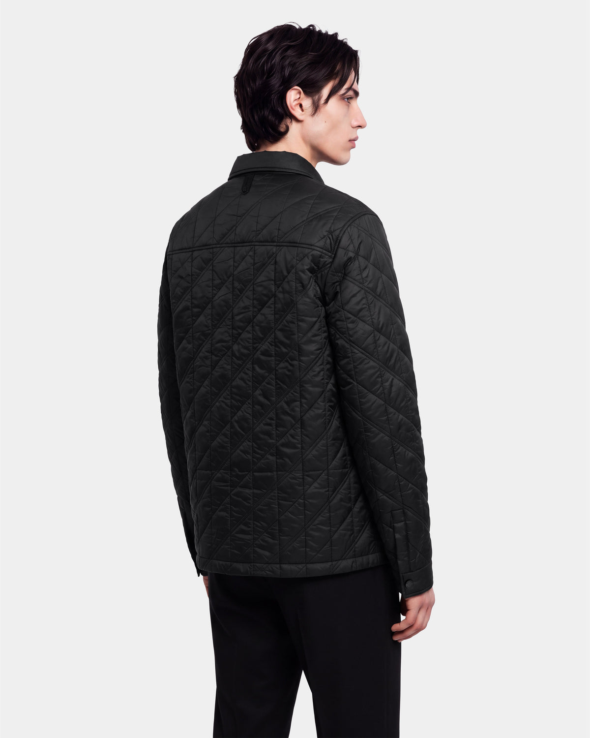 A person with dark hair is standing and facing away, looking to the side against a plain light background, wearing the Rudsak GRIFFIN - 6126758 Black quilted jacket and black pants.