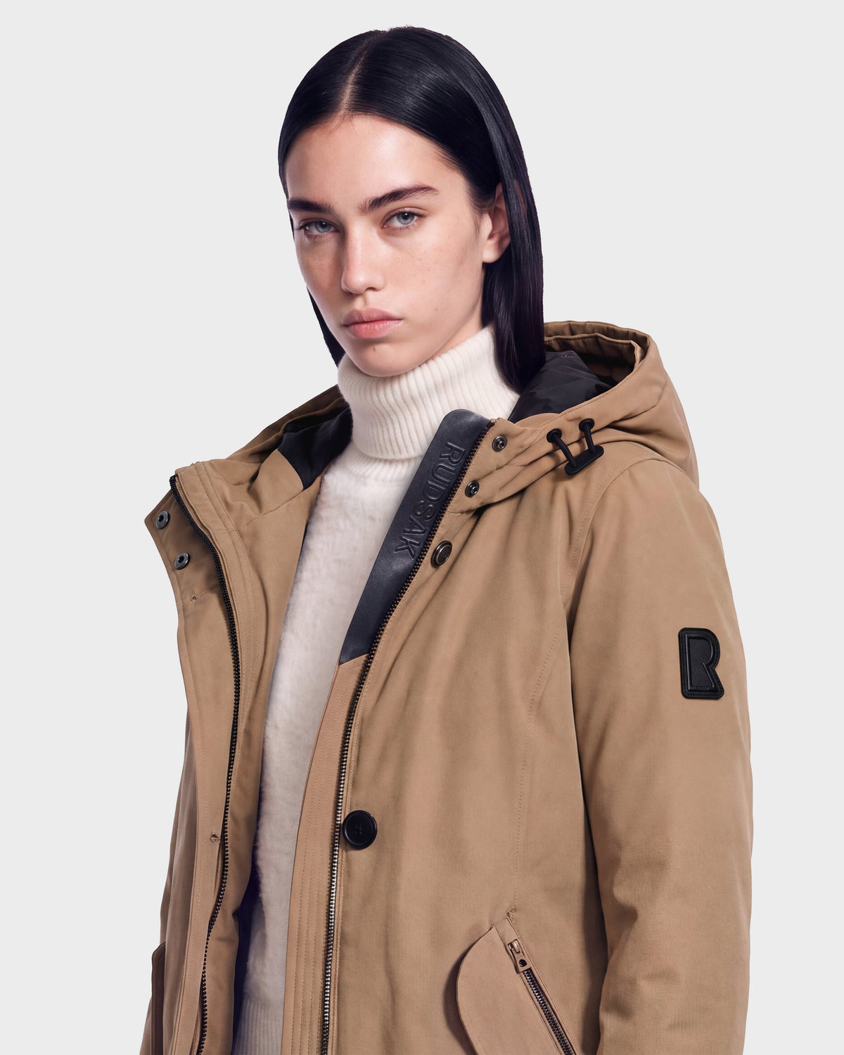 A woman with straight dark hair is wearing the Rudsak KEYSHIA - 8126517 Camel, an RDS-certified down women’s parka in a tan shade, layered over a white turtleneck sweater. She stands against a plain light background, looking directly at the camera with a neutral expression.