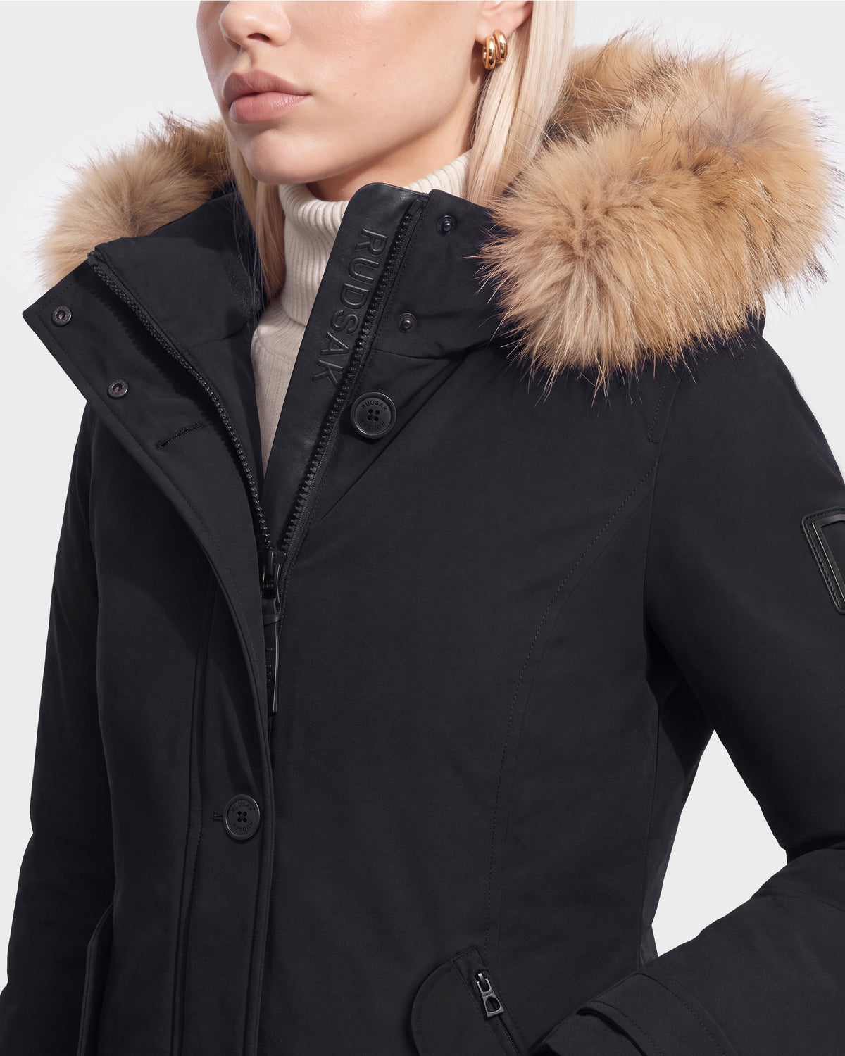 A woman wears the Rudsak KEYSHIA - 8126517 Black down parka with a fur-lined hood over a cream turtleneck, showing only the lower half of her face and upper body.