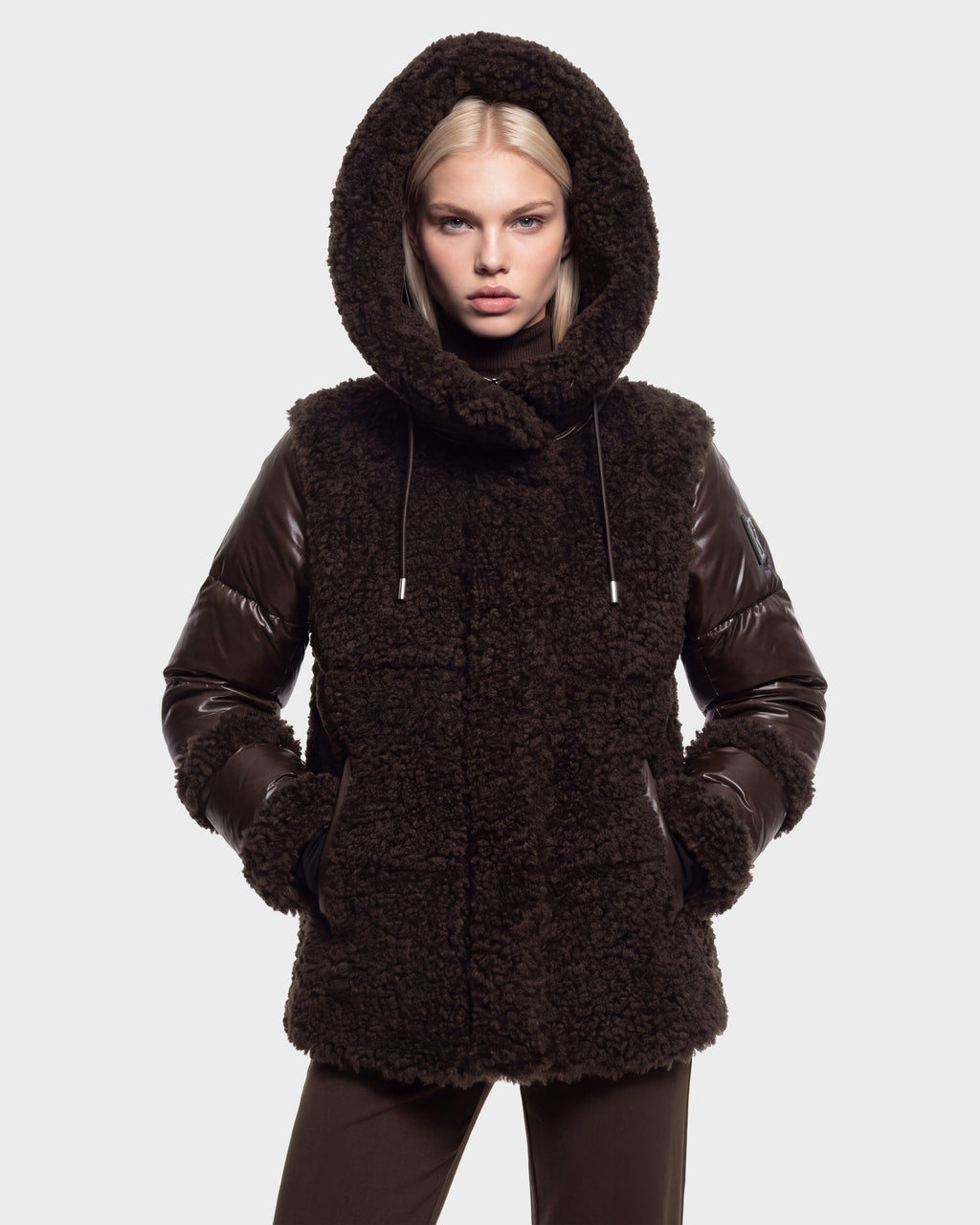 A person wearing the Rudsak MARISSA - 8126728 Espresso coat, a dark brown textured winter outerwear piece with leather sleeves and a hood, stands against a plain light gray background with hands in pockets, looking directly at the camera.