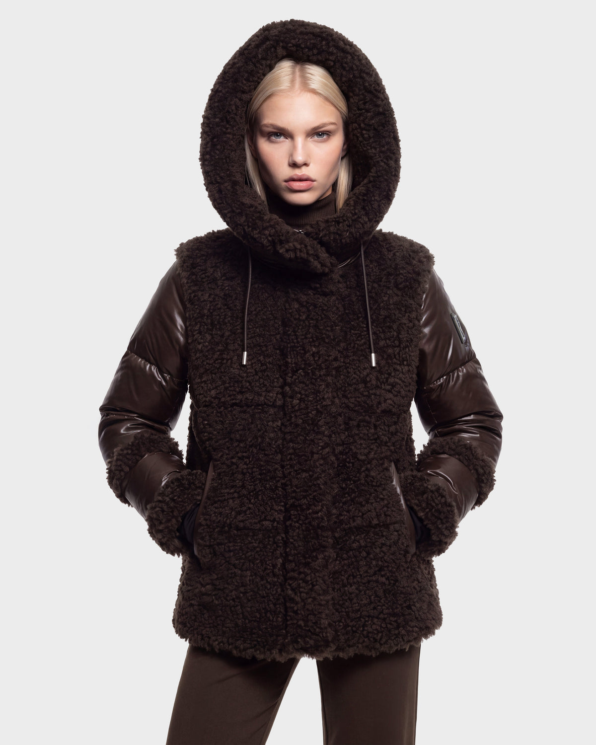A person wearing the Rudsak MARISSA - 8126728 Espresso coat, a dark brown textured winter outerwear piece with leather sleeves and a hood, stands against a plain light gray background with hands in pockets, looking directly at the camera.
