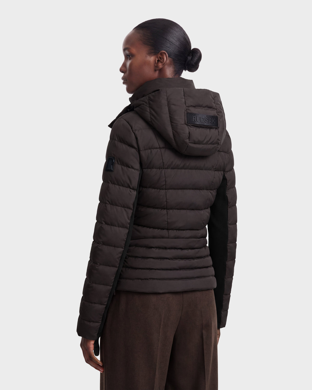 A woman with tied-back hair, viewed from the back and slightly to the side against a plain light background, is wearing the Rudsak MELISSA - 8124824 lightweight down puffer jacket with a hood in Espresso/Black.