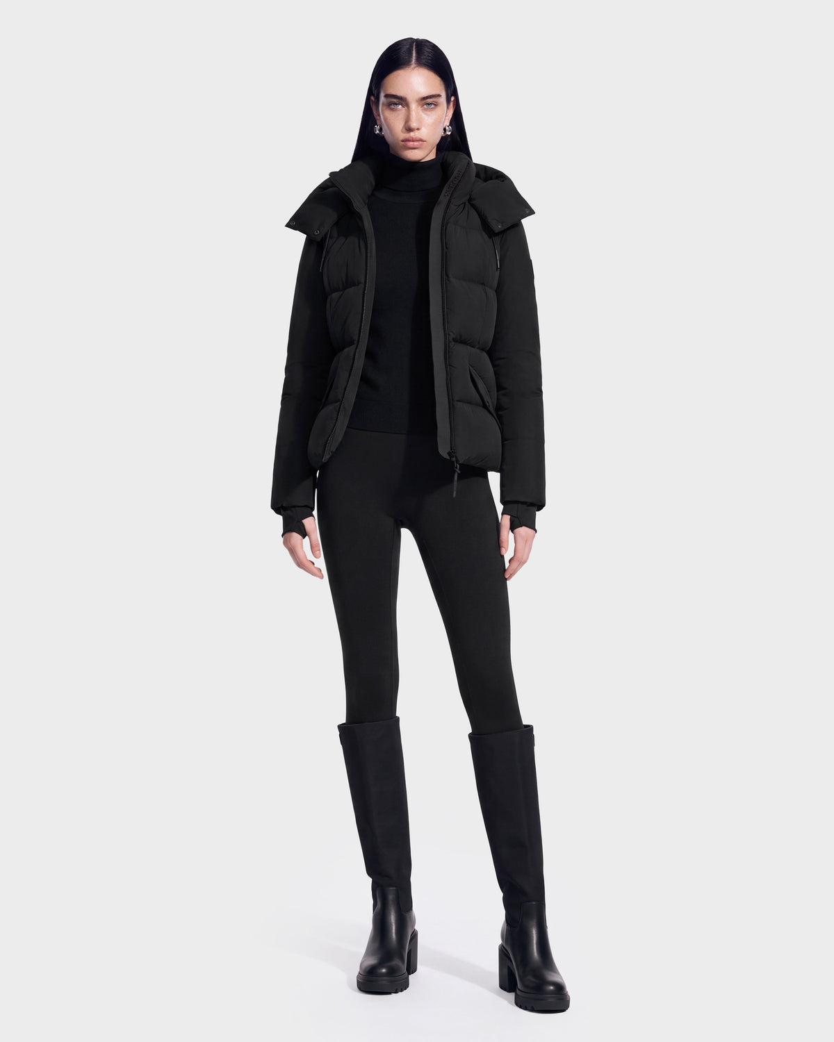 A person stands facing forward, wearing the Rudsak ADELINE - 8124544 Black women’s down puffer jacket made with RDS-certified down, paired with a black turtleneck, black leggings, and tall black boots against a plain white background.