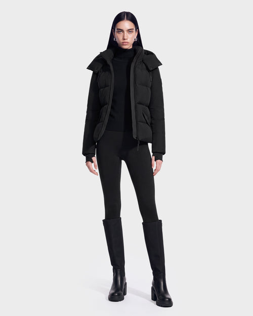 A person stands facing forward, wearing the Rudsak ADELINE - 8124544 Black women’s down puffer jacket made with RDS-certified down, paired with a black turtleneck, black leggings, and tall black boots against a plain white background.