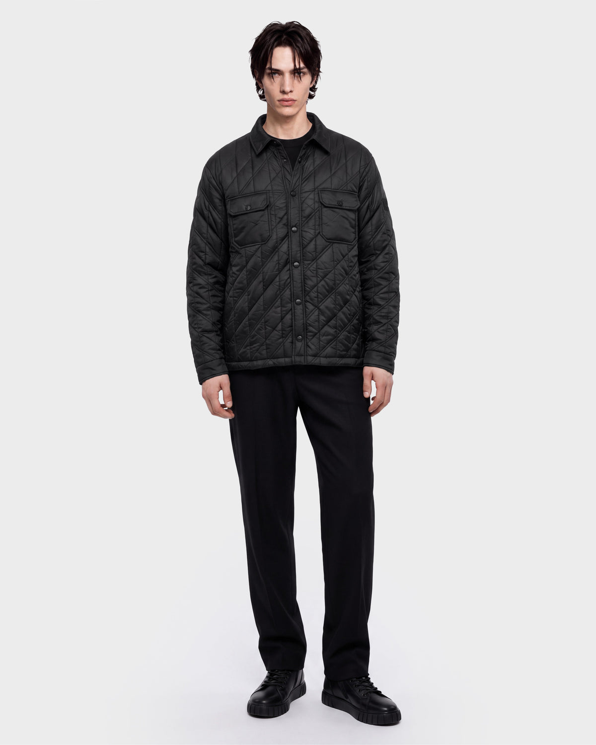 A person stands facing forward wearing the Rudsak GRIFFIN - 6126758 Black quilted jacket, paired with black pants and black shoes, against a plain white background.