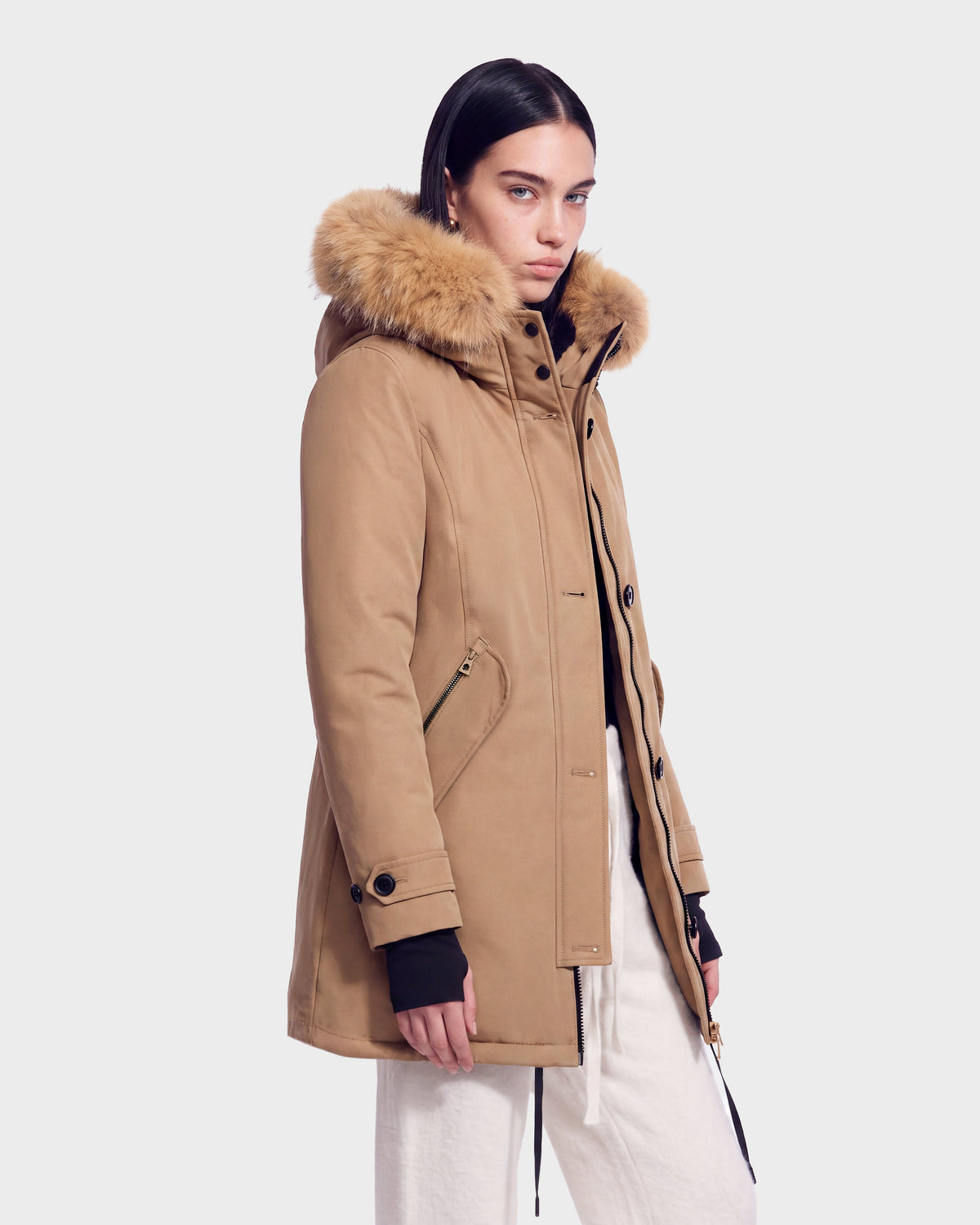 A woman wearing the Rudsak KEYSHIA - 8126517 Camel parka, crafted from RDS-certified down with a fur-lined hood and paired with white pants, stands against a plain light background, gazing at the camera with a neutral expression.