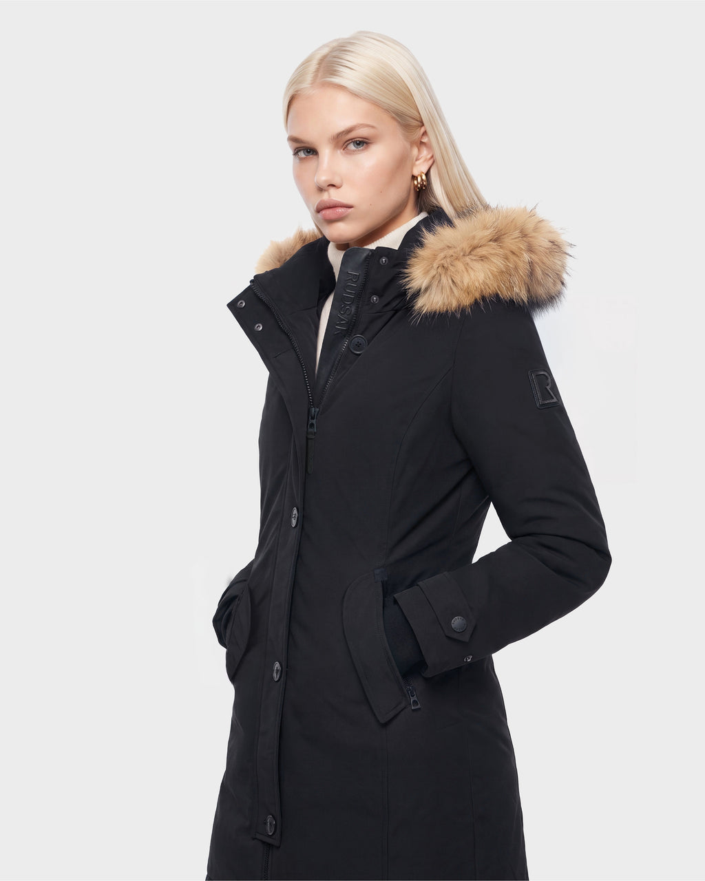 A woman with straight blonde hair wears the Rudsak KEYSHIA - 8126517 Black women’s down parka with a fur-lined hood, standing with hands in pockets against a plain white background.