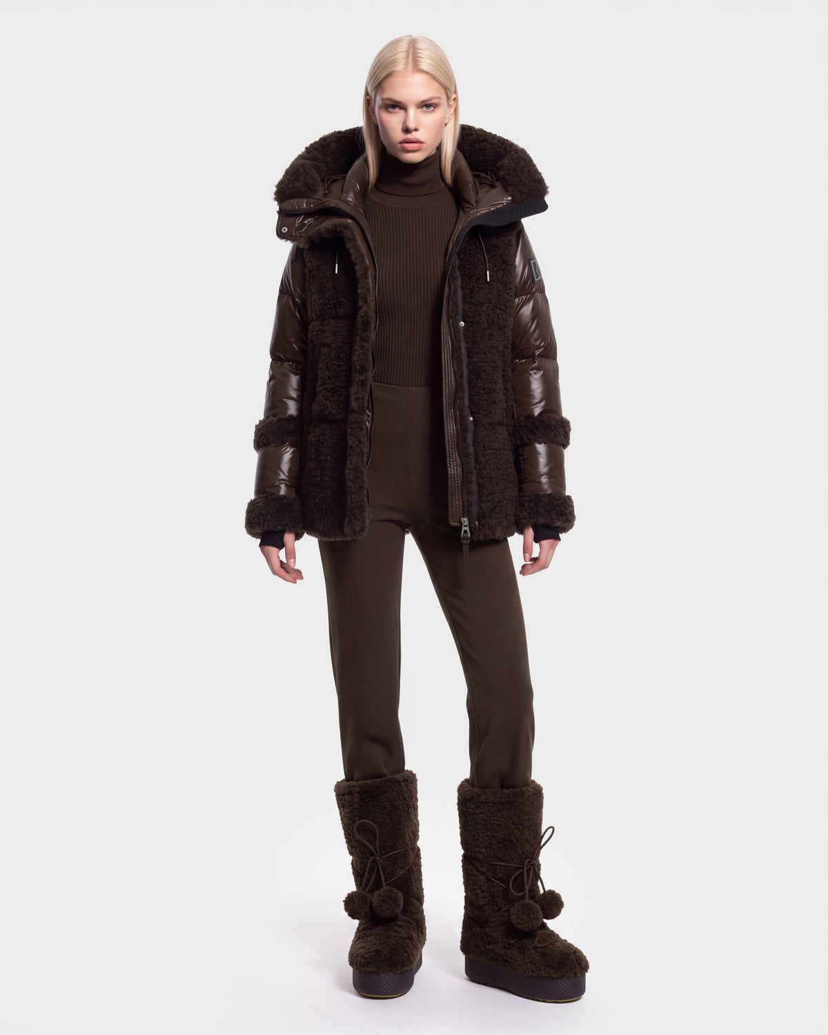 A person stands facing forward, wearing the Rudsak MARISSA - 8126728 Espresso dark brown down insulated puffer coat with fur trim, paired with matching brown pants and large brown fur boots—perfect winter outerwear. The background is plain white.
