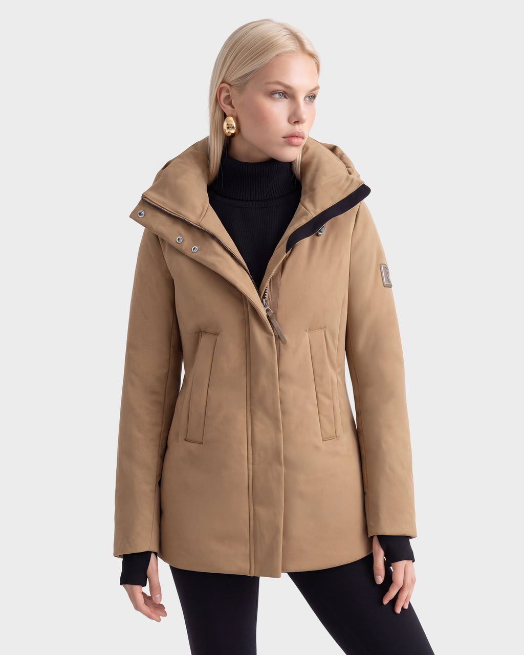 A person with blonde hair wears the Rudsak OPAL - 8126618 Camel parka, black turtleneck, and black pants, standing against a plain white background and looking to the side.