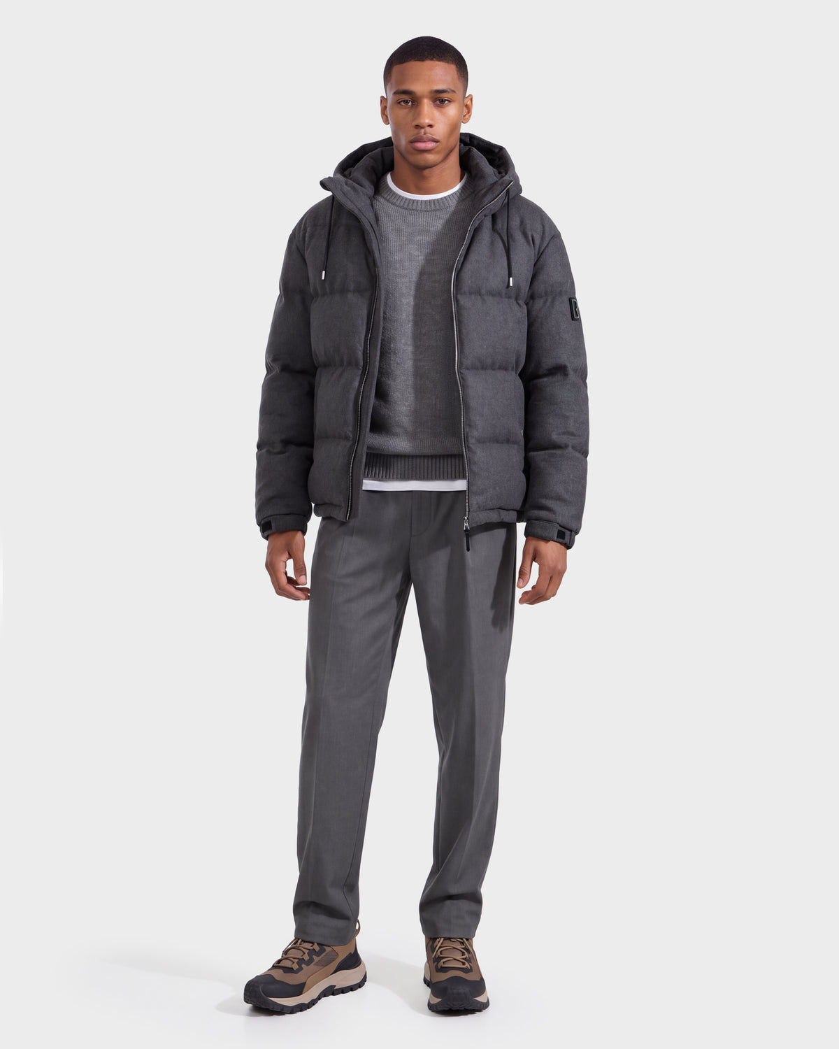 A man wearing the Rudsak STERLING W - 6126760 Heather Charcoal puffer jacket over a gray sweater and shirt, paired with gray pants and brown sneakers, stands facing forward against a plain white background.