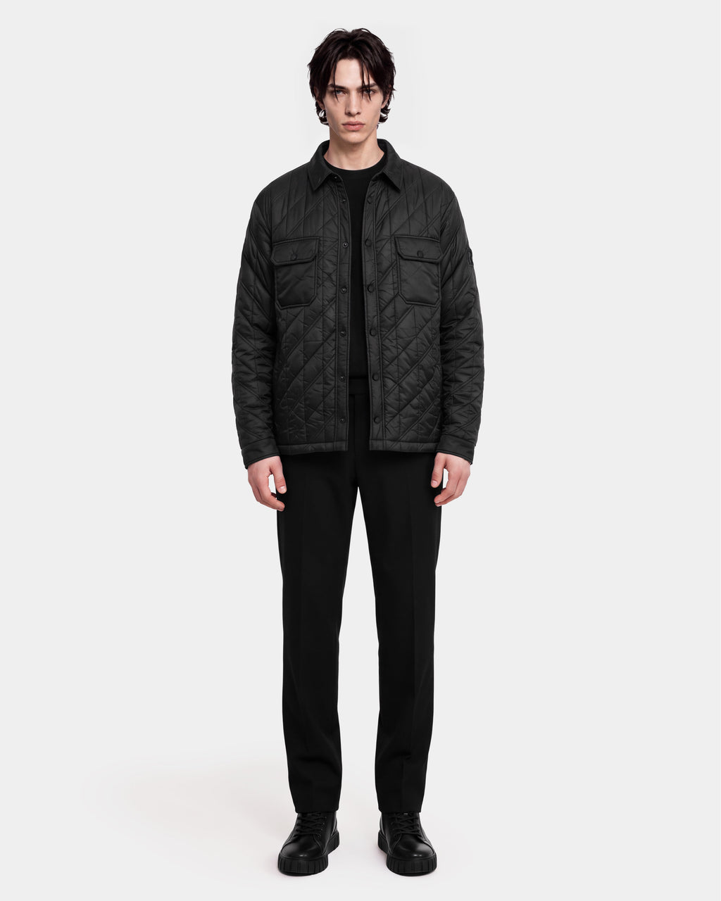 A person stands facing forward against a plain white background, wearing the Rudsak GRIFFIN - 6126758 Black quilted jacket, paired with a black shirt, black pants, and black shoes.